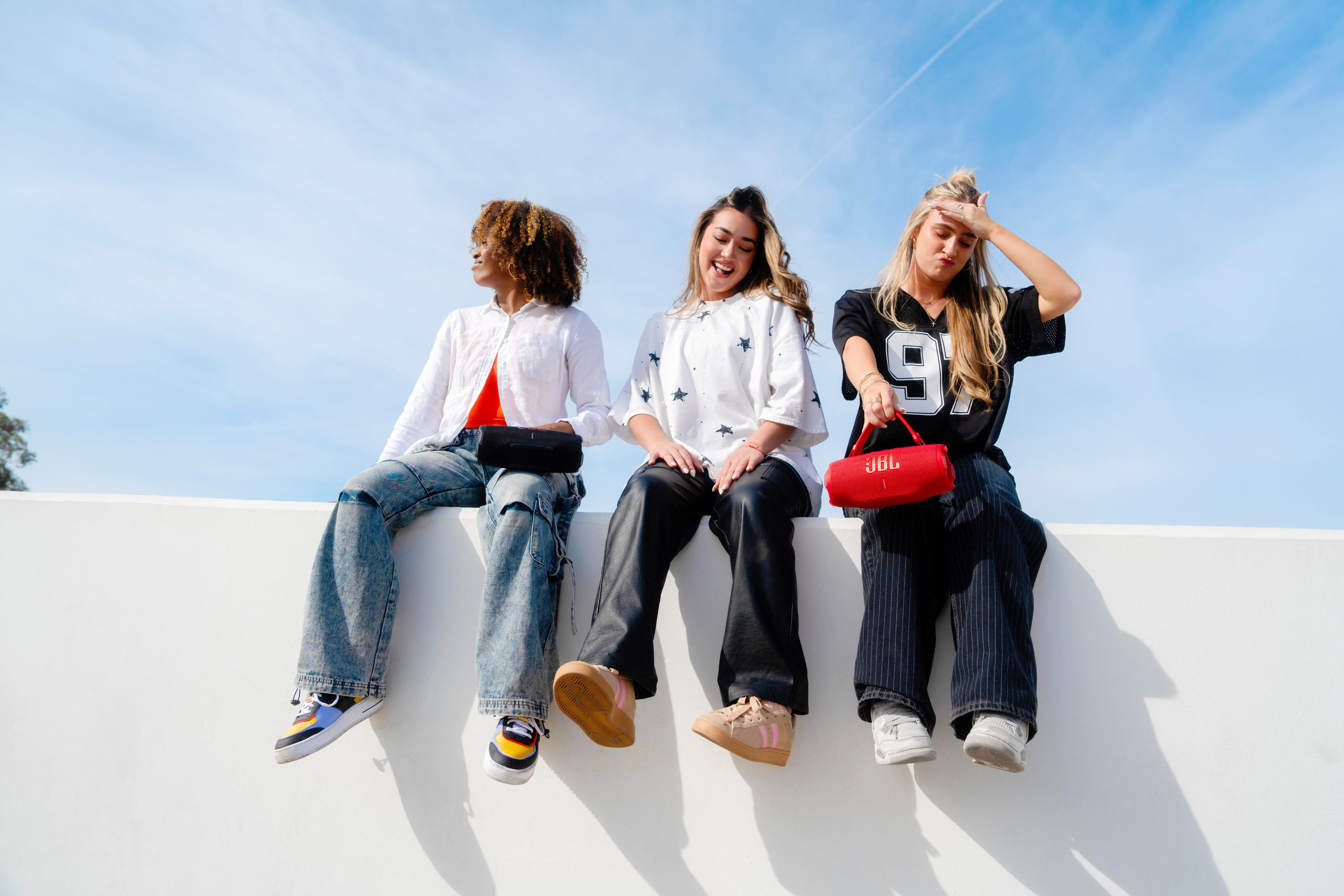 Drei Frauen sitzen auf einer Mauer, halten eine Tasche und einen Lautsprecher, mit blauem Himmel.