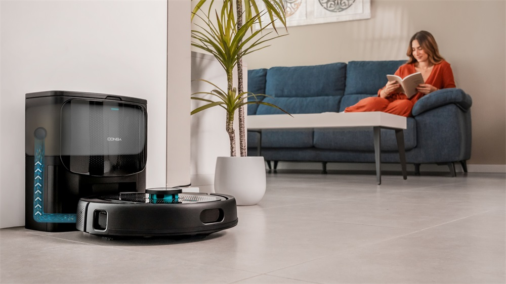 Robot porszívó és töltőállomás a csempén. Nő olvas a kék kanapén a háttérben.