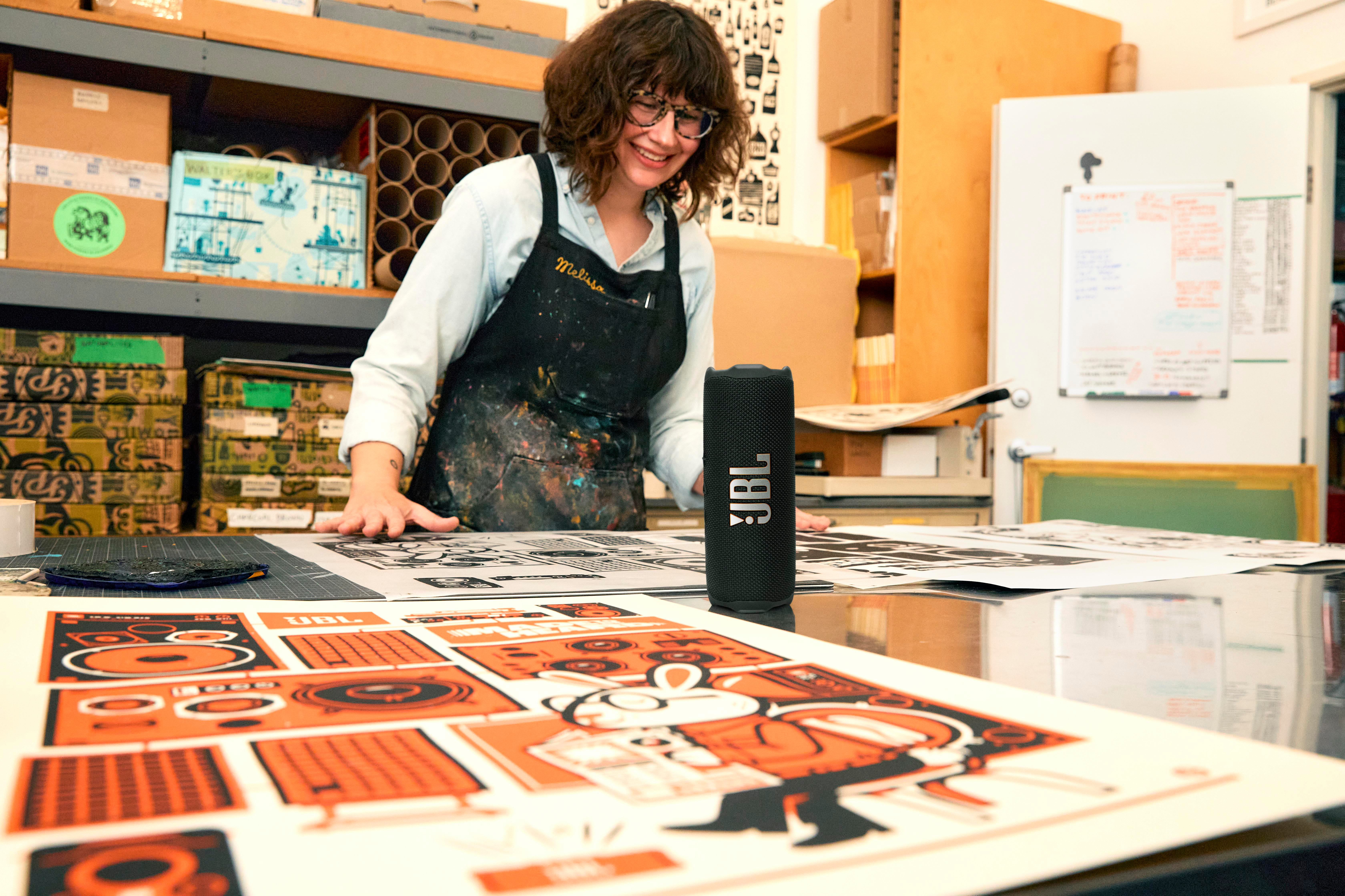 Frau in einem Kunstatelier mit einem Lautsprecher. Sie lächelt, umgeben von Kunstwerken.