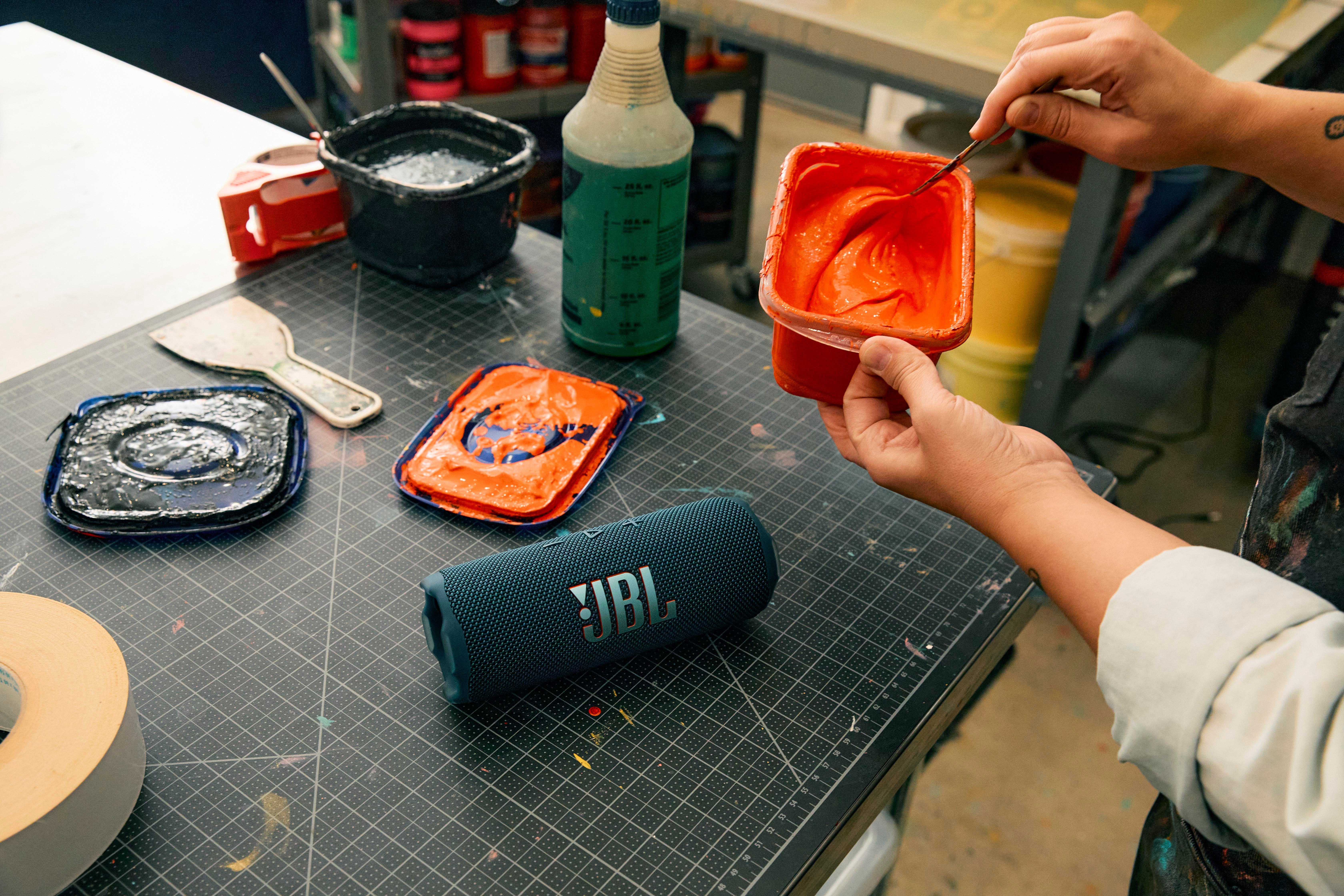 Hände mischen orange Farbe in einem Behälter. JBL-Lautsprecher und Malutensilien vorhanden.
