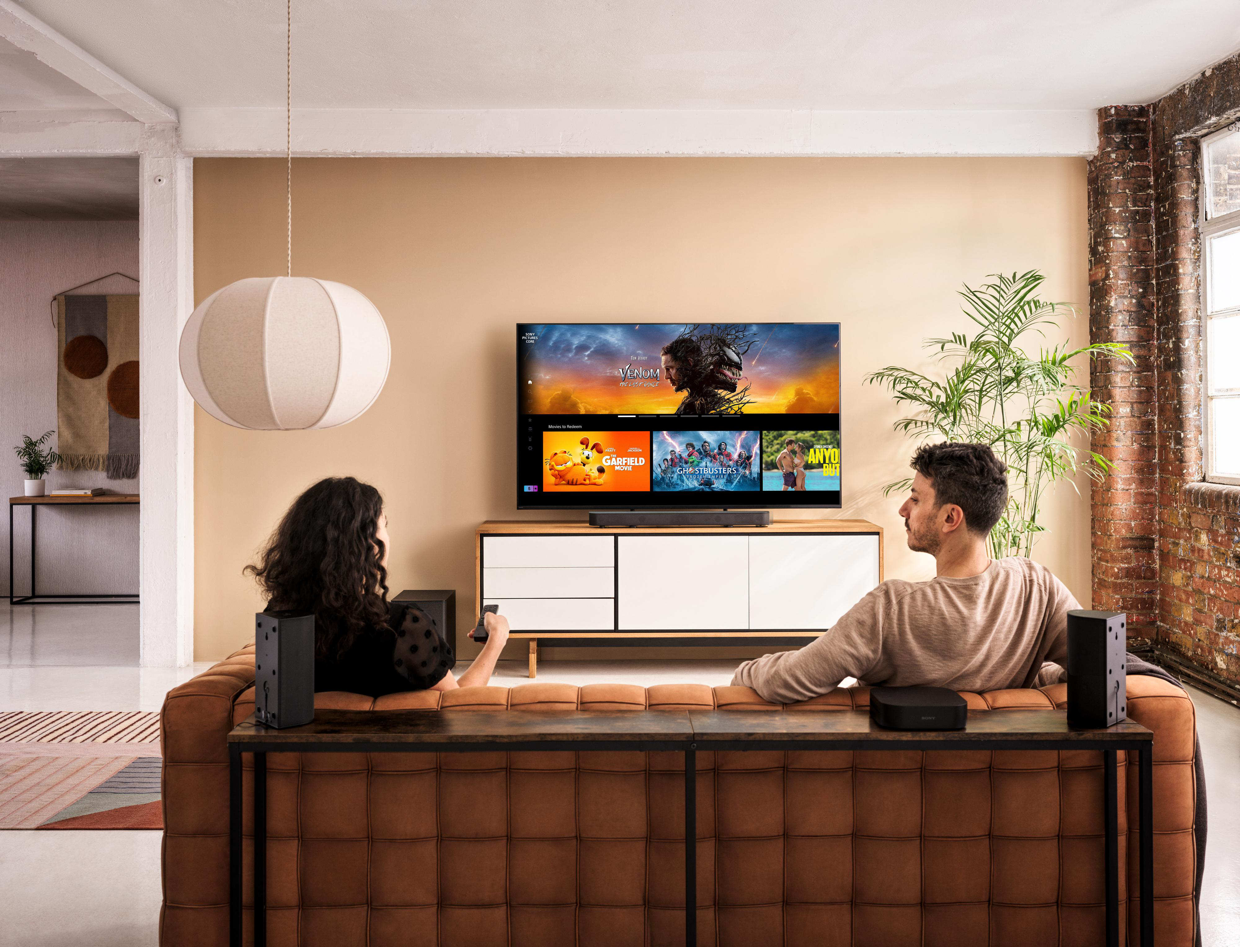 Ein Wohnzimmer mit Fernseher, zwei sitzenden Personen und Lautsprechern auf beiden Seiten.