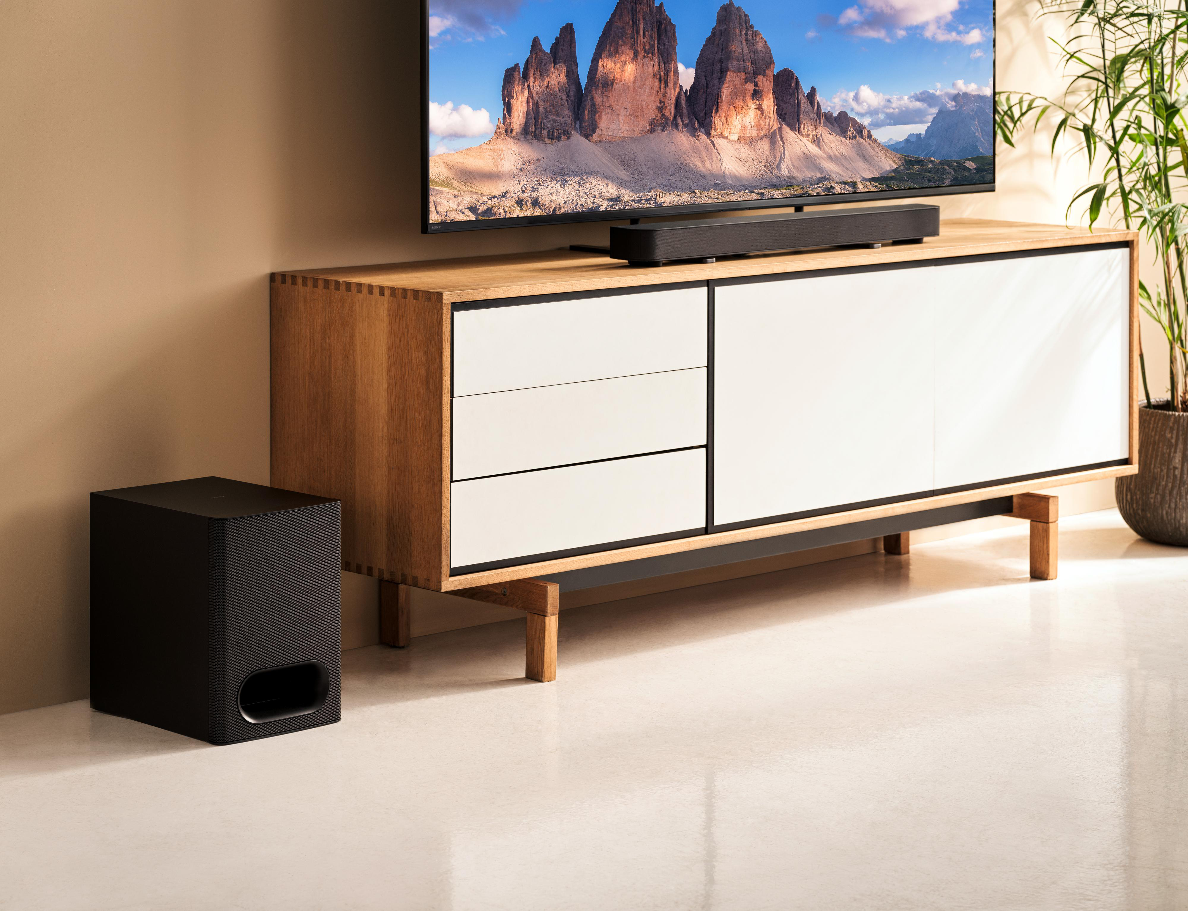 Wohnzimmer mit Fernseher auf Holzschrank, Soundbar und schwarzem Subwoofer.