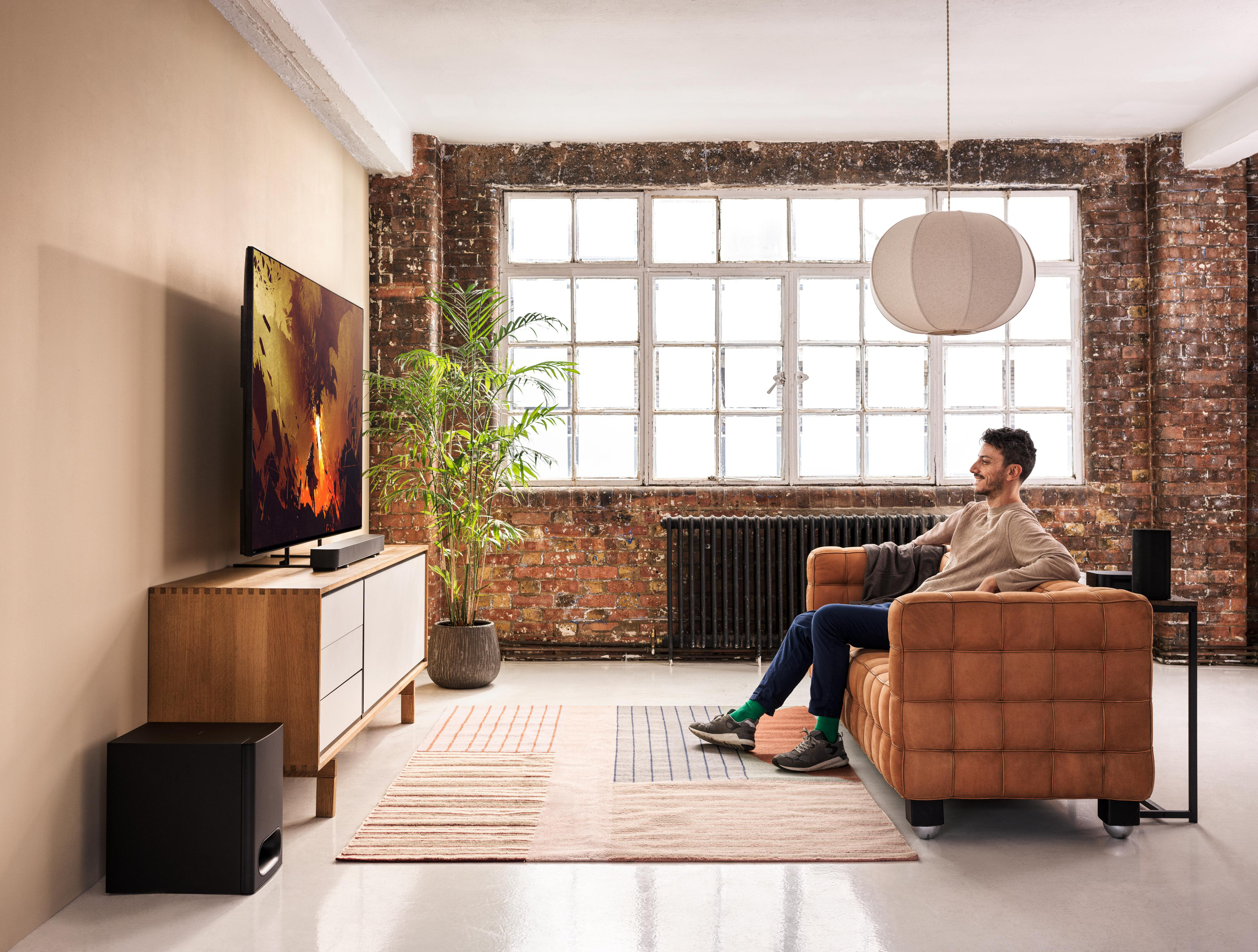 Mann schaut Fernsehen mit Soundbar und Subwoofer. Braune Couch und Backsteinmauer.