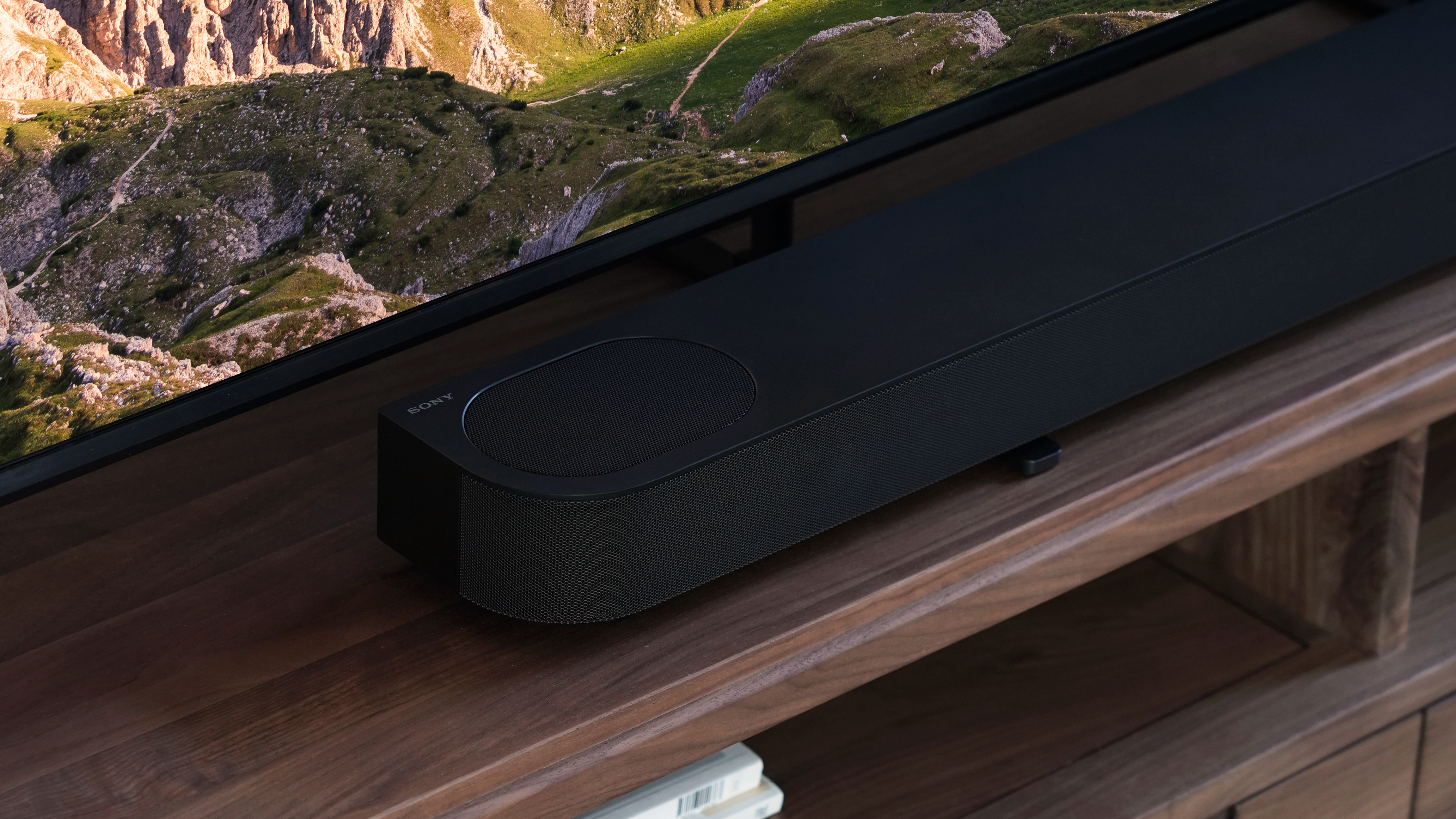 Eine schwarze Soundbar auf einer Holzoberfläche vor einem Fernseher. Der Lautsprecher hat ein Holzfinish und ein Logo.