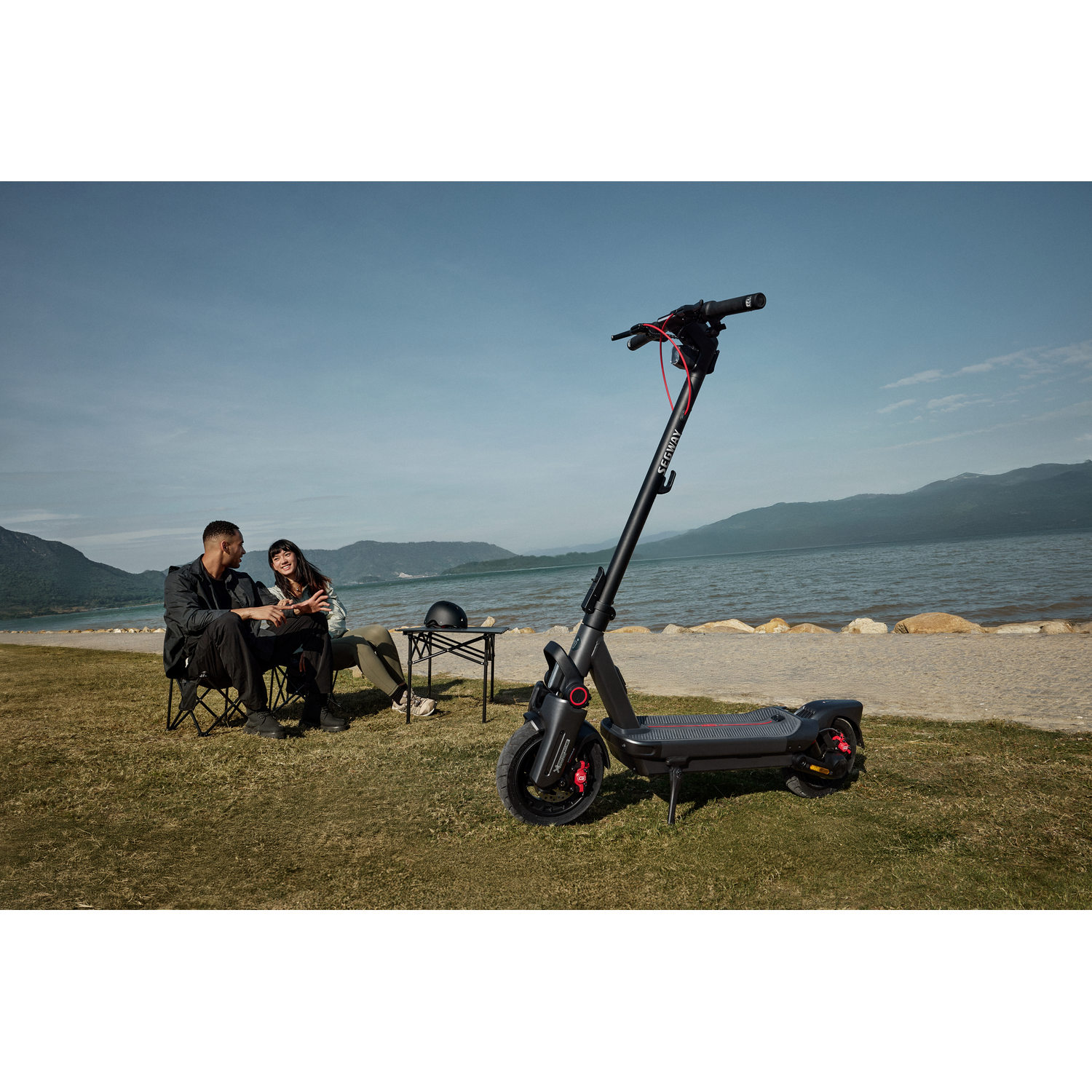 Koppel zit op gras bij een elektrische scooter, bergen en meer op de achtergrond, blauwe lucht.