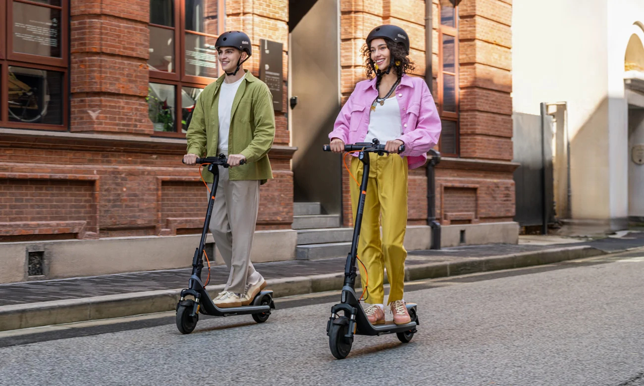 Dwie osoby jadące na hulajnogach elektrycznych na ulicy miejskiej z budynkami w tle.