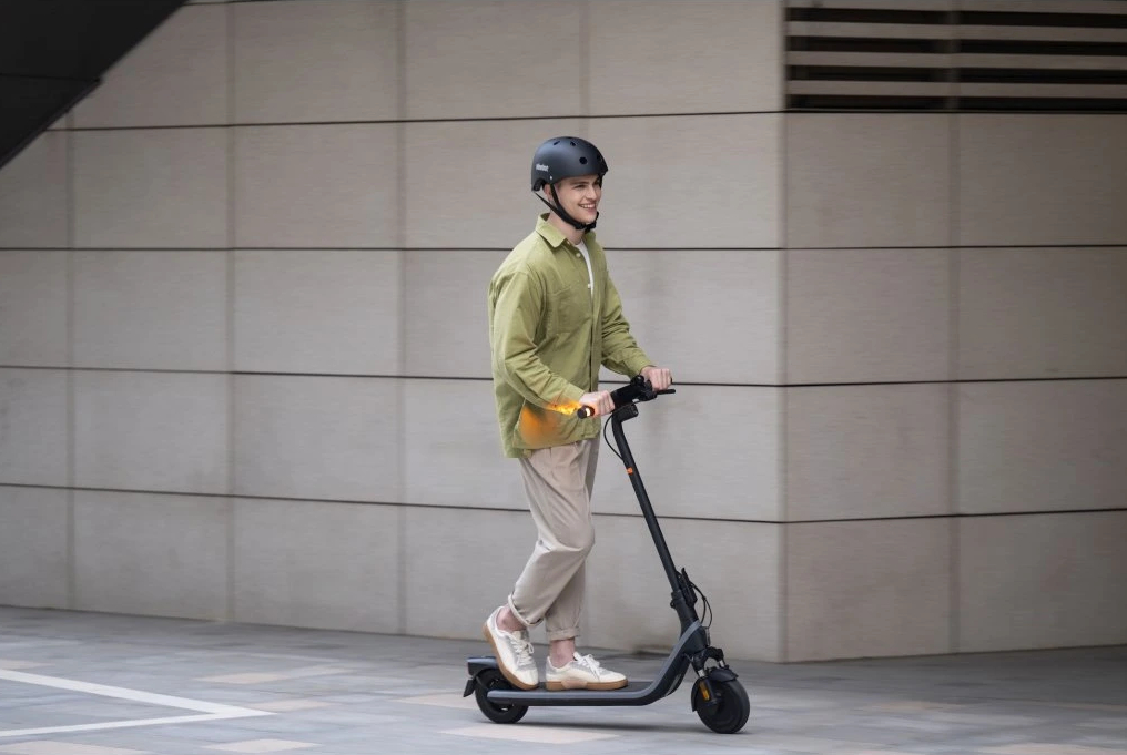 Mężczyzna jedzie hulajnogą elektryczną po utwardzonym terenie. Uśmiecha się i ma na sobie kask.