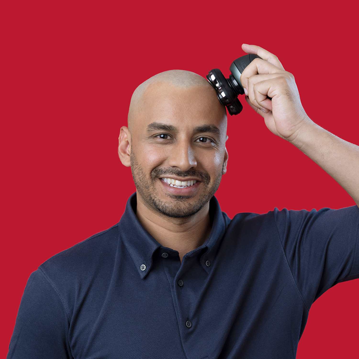 Een man scheert zijn hoofd met een zwart elektrisch scheerapparaat. Hij glimlacht, met een rode achtergrond.