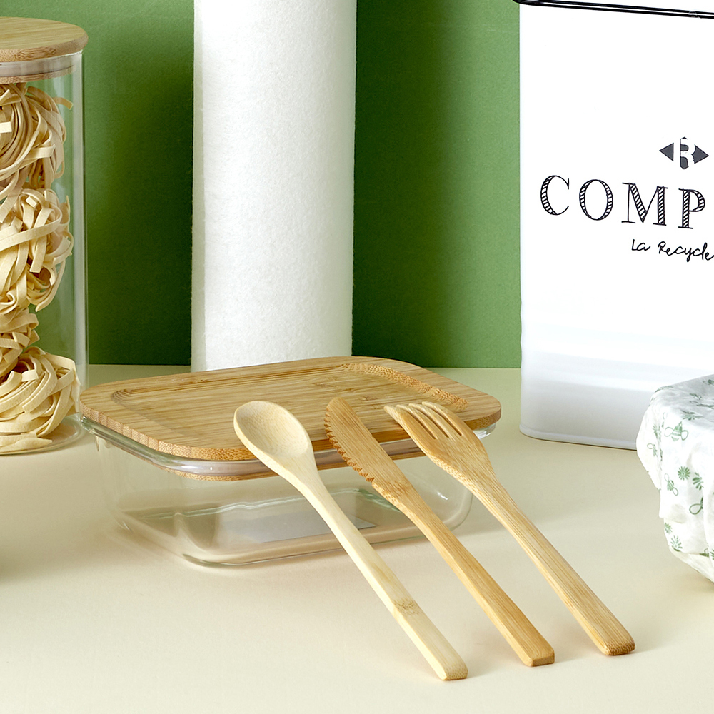 Un recipiente de vidrio, cuchara, cuchillo y tenedor de madera sobre una mesa, con un contenedor blanco.