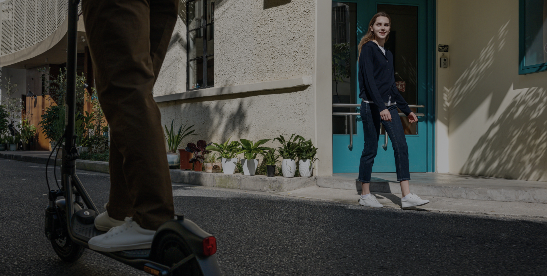 Osoba na hulajnodze mija kobietę idącą ulicą. W tle budynki i rośliny.