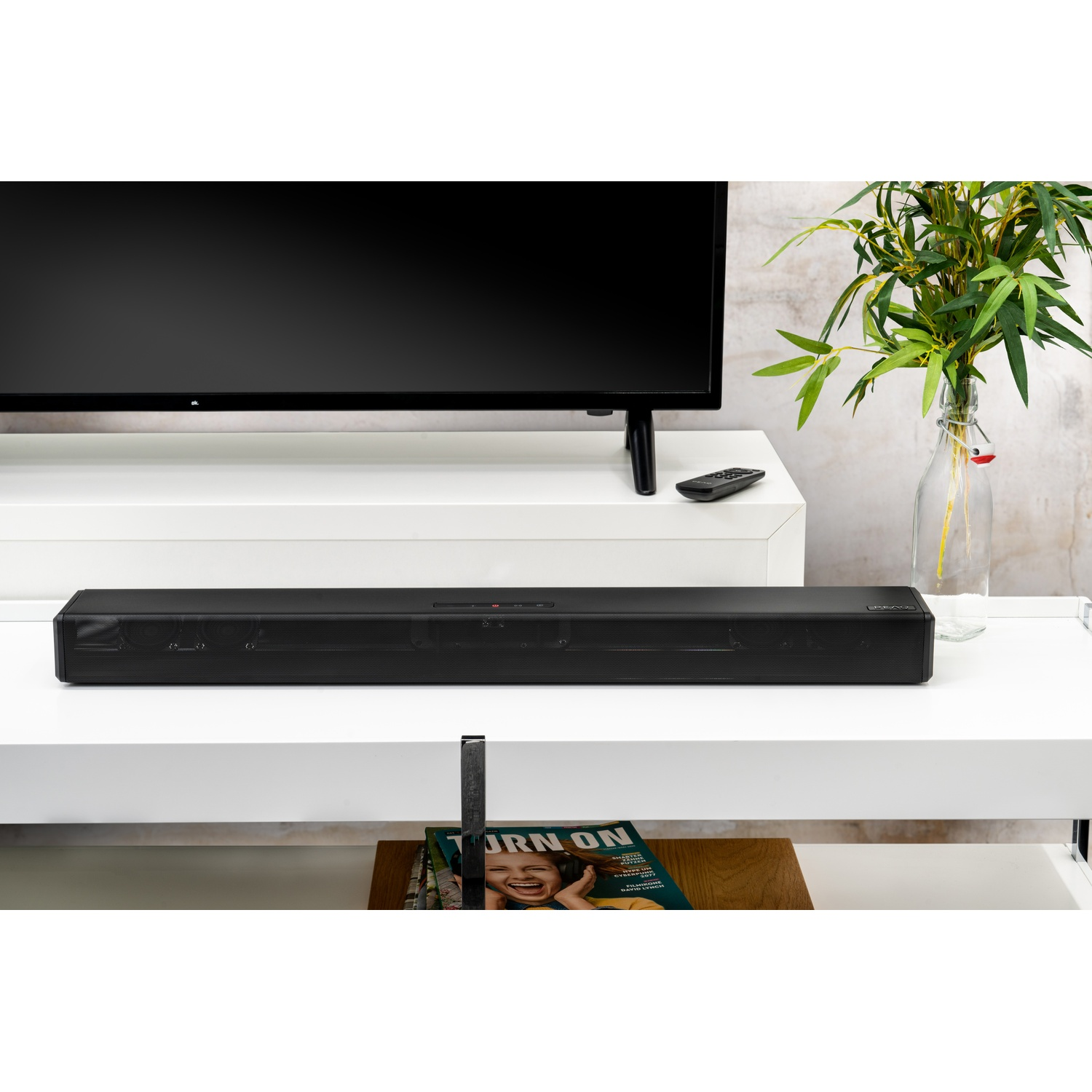 Zwarte soundbar op witte plank, met tv erboven. Vaas met planten rechts, afstandsbediening.