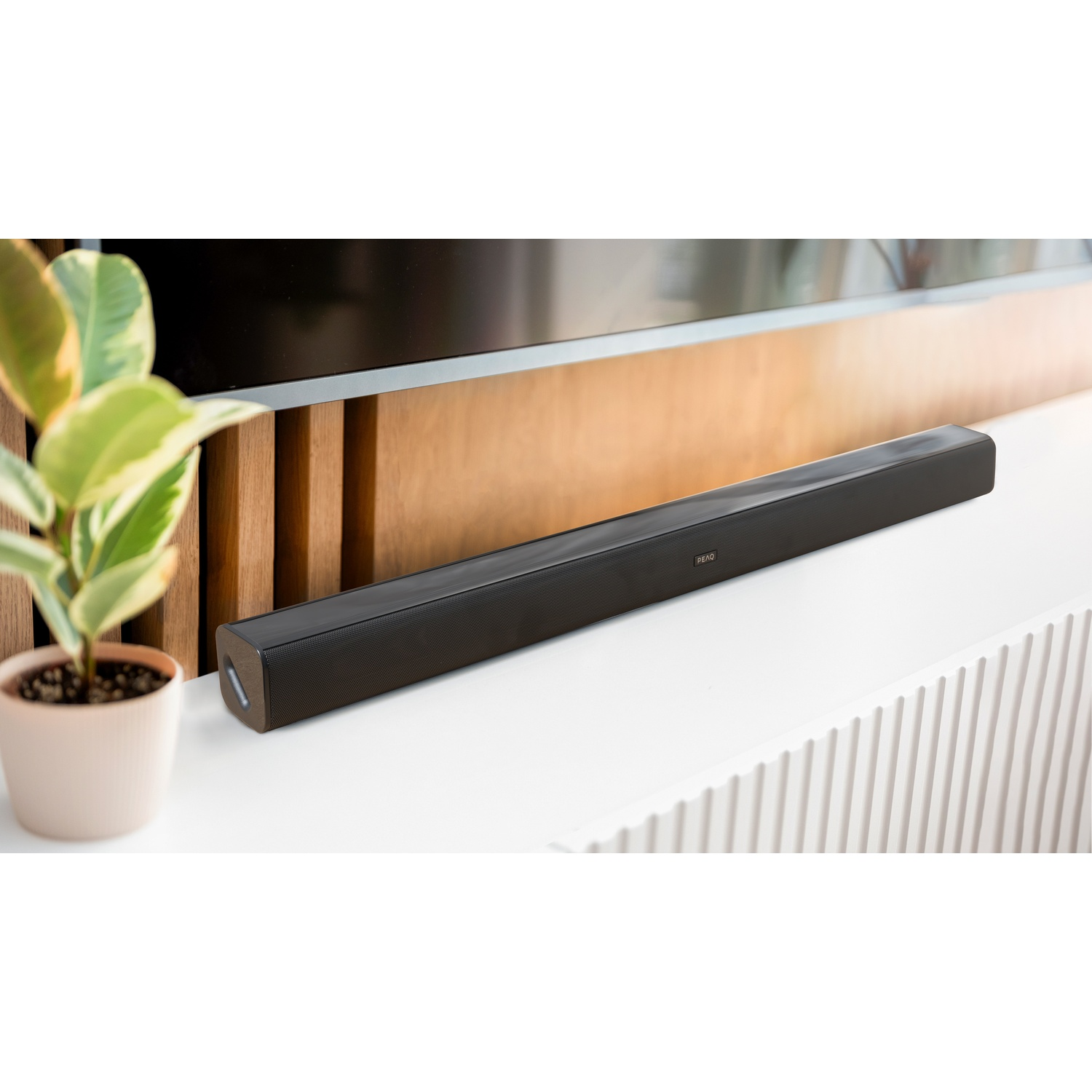 Zwarte soundbar op een wit oppervlak, met een plant en een tv op de achtergrond.