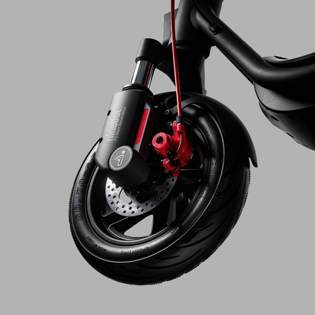 Zbliżenie koła skutera, czarna opona, czerwony hamulec i czerwony kabel, na szarym tle.