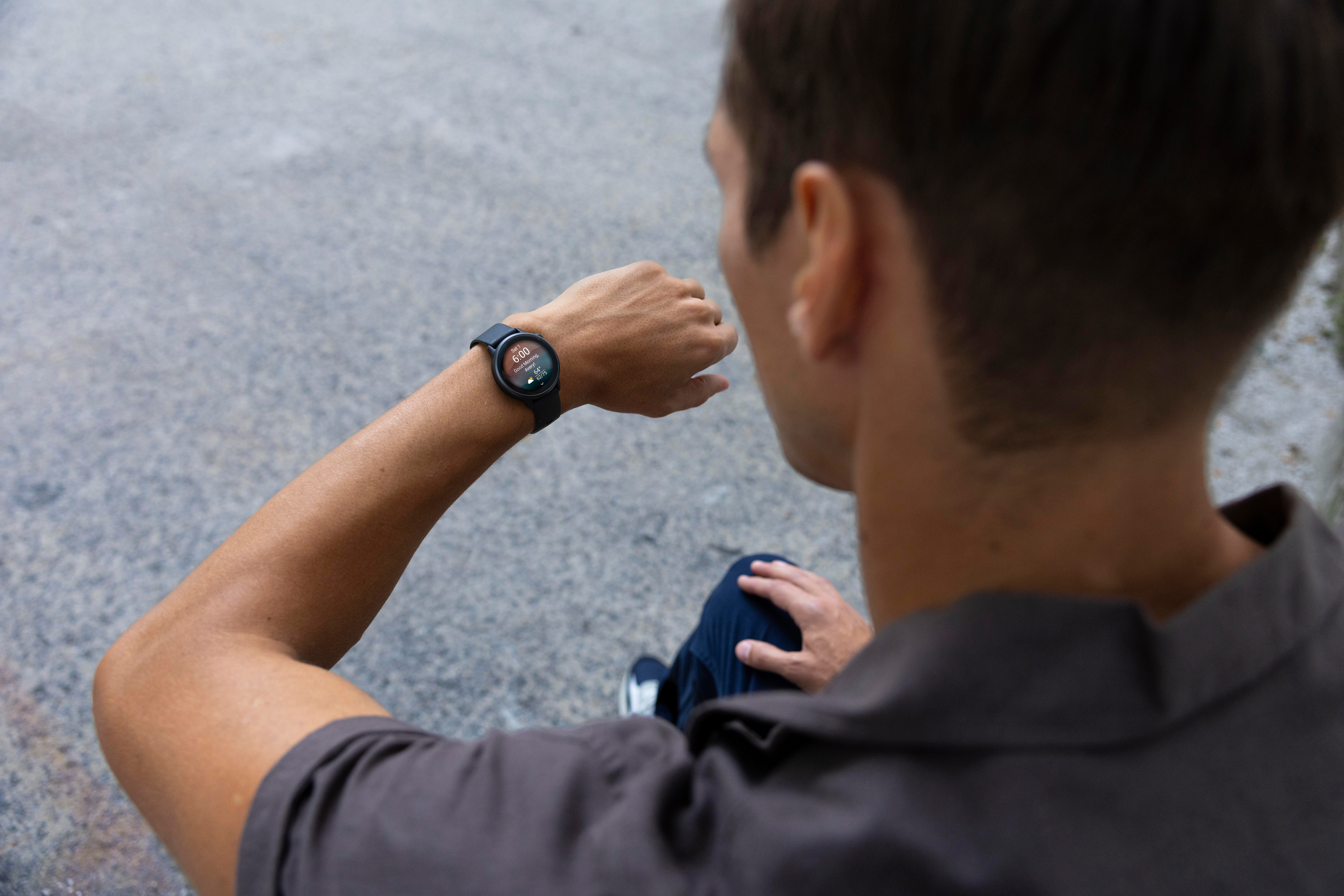 Eine Person betrachtet die Smartwatch an ihrem Handgelenk. Die Uhr zeigt Zeit und Daten an. Der Hintergrund ist eine verschwommene Pflasteroberfläche.