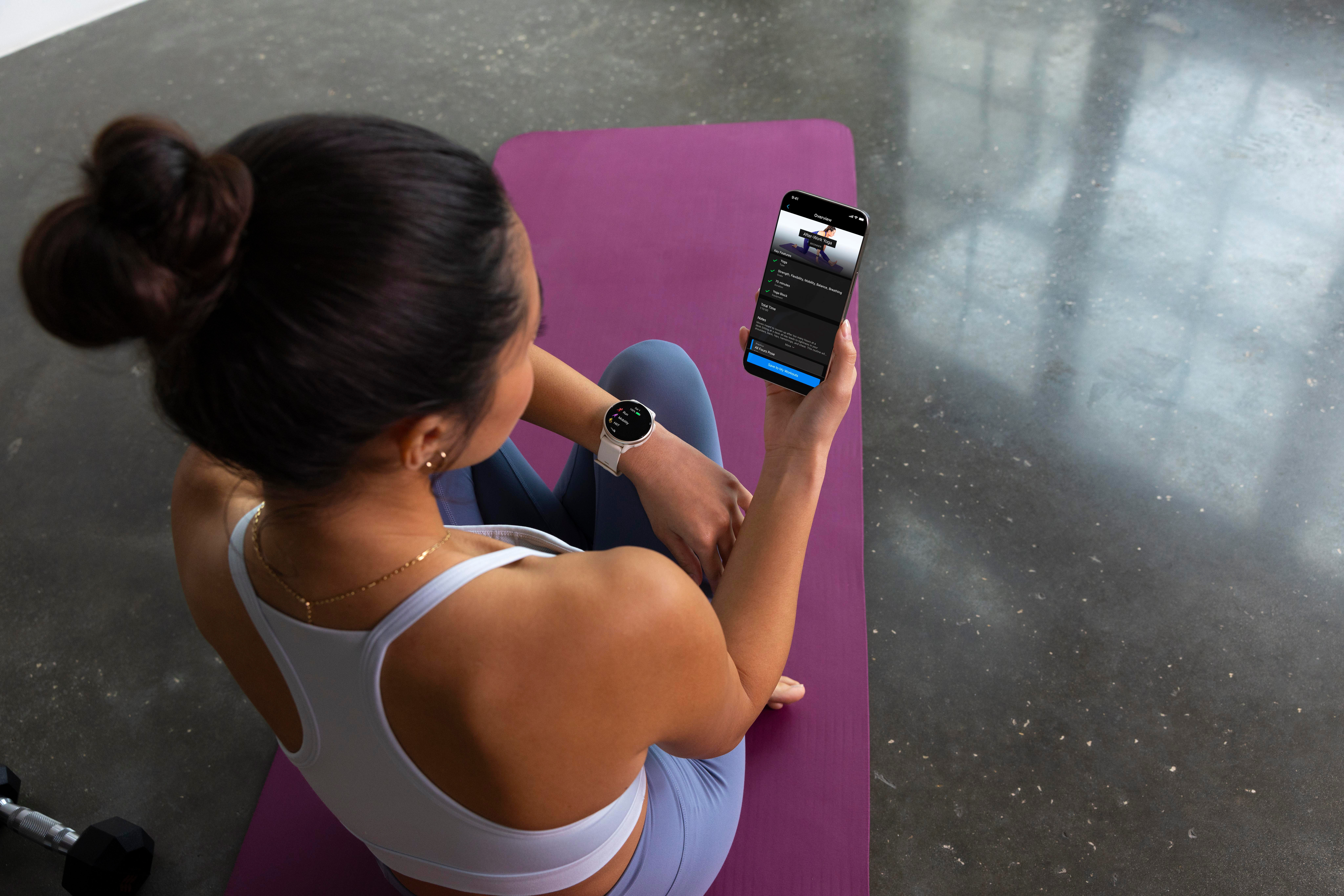 Frau sitzt auf einer Yogamatte und hält ein Telefon mit einer Workout-App in der Hand. Sie trägt eine Smartwatch. Im Hintergrund ist ein grauer Boden zu sehen.
