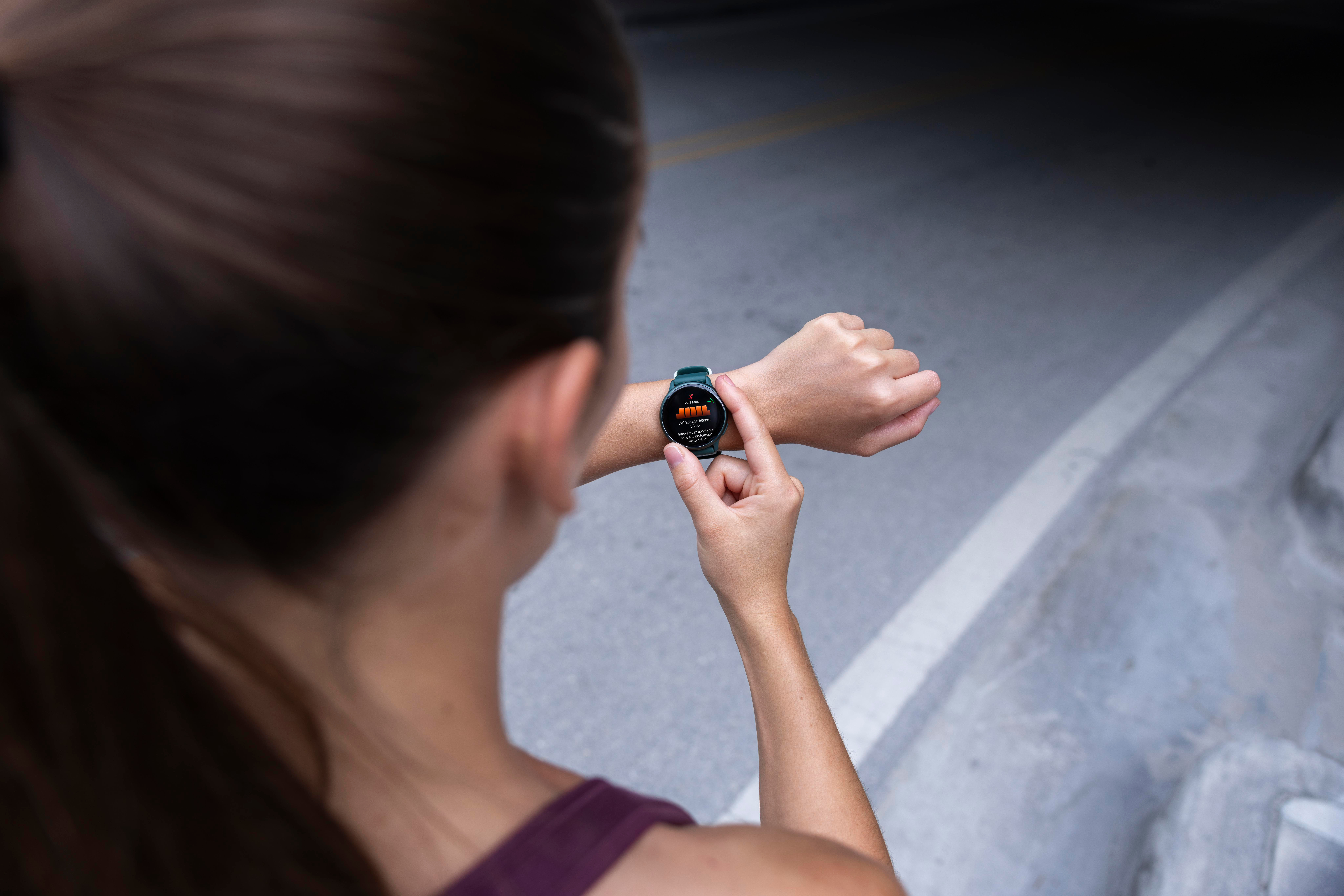 Eine Frau überprüft eine Smartwatch mit Trainingsdaten. Im Hintergrund sind Straßenmarkierungen zu sehen.