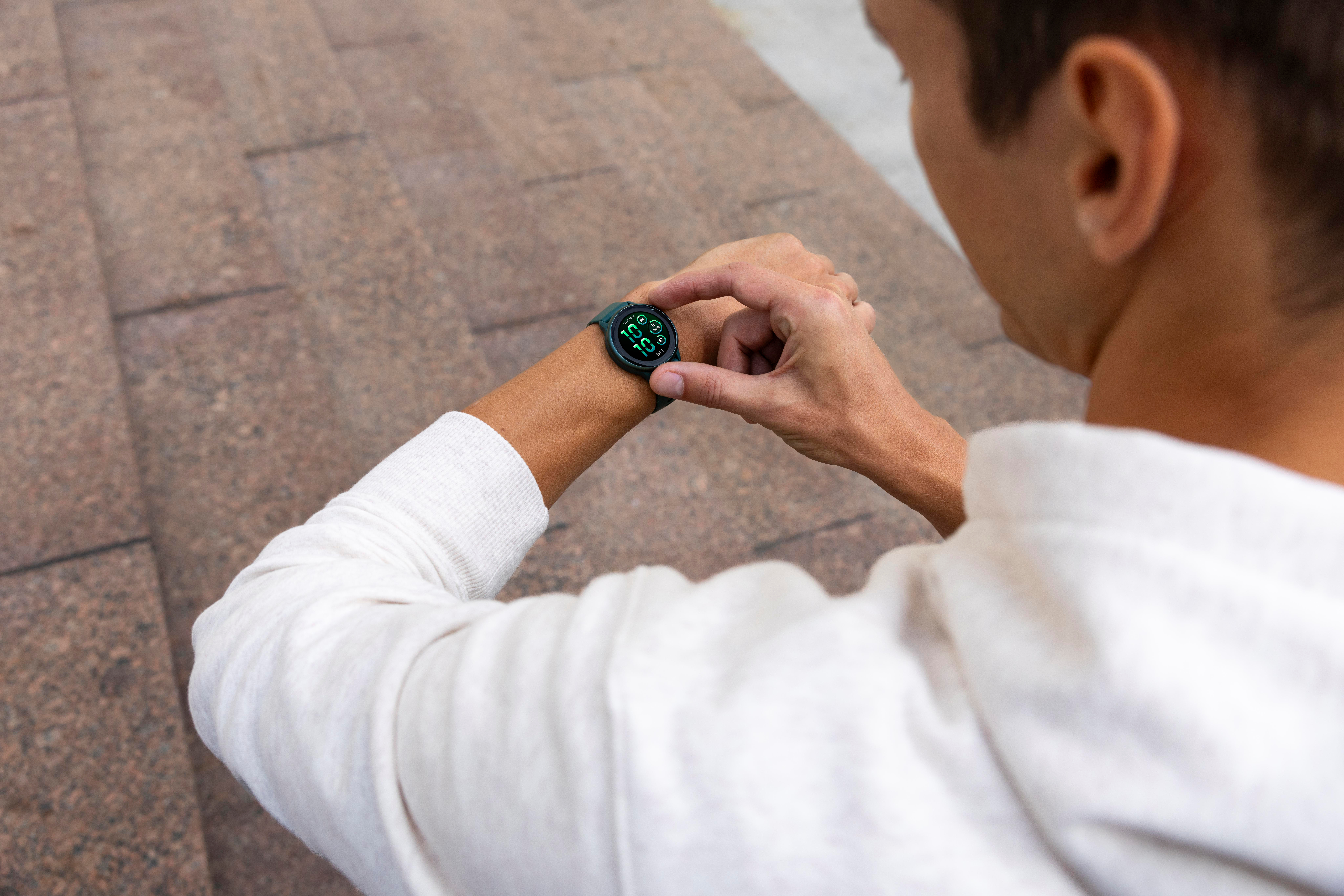 Person, die eine grüne Smartwatch trägt und sie betrachtet. Trägt einen weißen Pullover. Im Hintergrund ist Steinpflaster zu sehen.