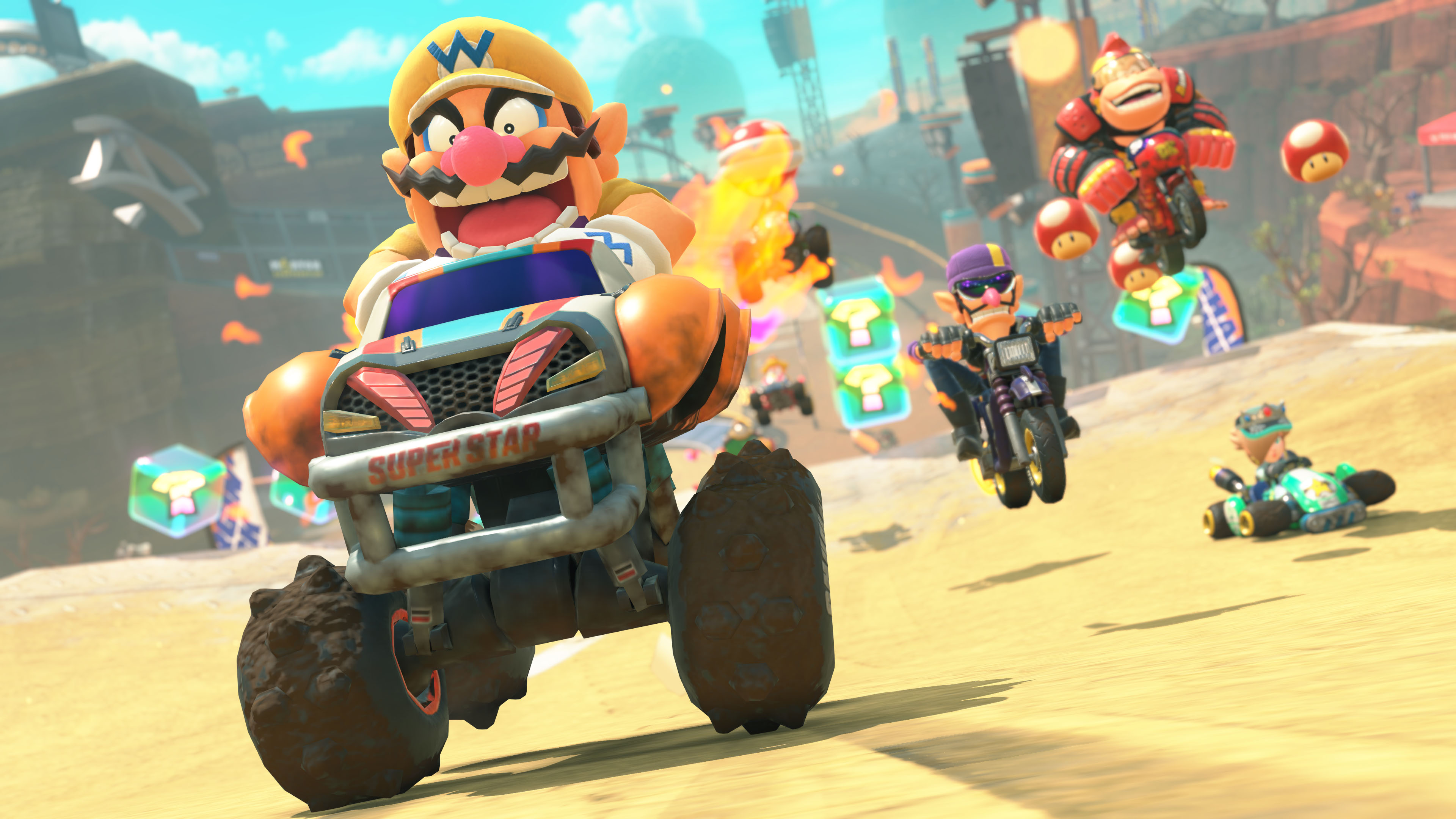 Mario Kart-Szene mit Wario in einem Monstertruck-Kart. Andere Charaktere rasen auf einer Wüstenstrecke.