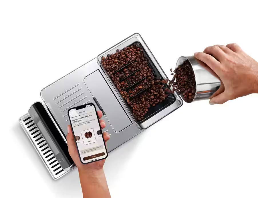 Osoba wsypująca ziarna kawy do ekspresu. Telefon pokazuje opcje wyboru kawy.