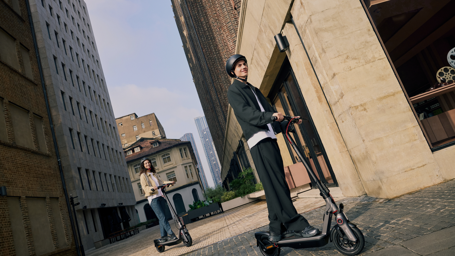 Dwie osoby na skuterach jadą ulicą miejską z wysokimi budynkami.