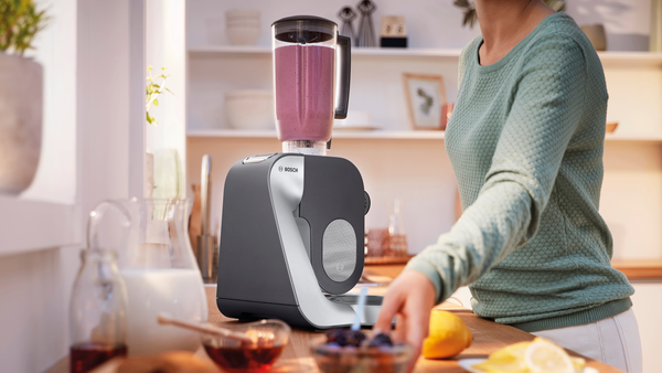 Kadın, blender ile smoothie yapıyor. Tezgahta malzemeler ve limon var. Arkada pencere.