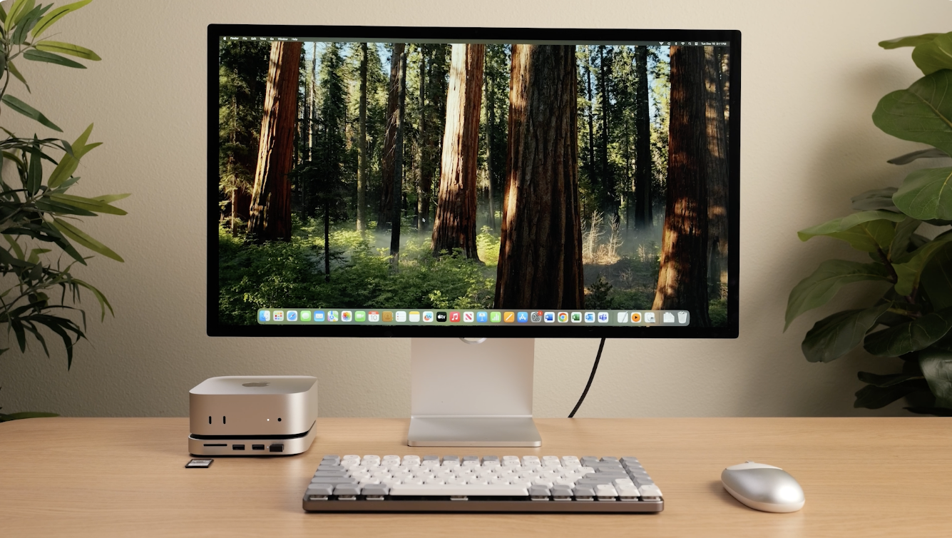 Schreibtisch mit Computerbildschirm, Tastatur, Maus und kleinem silbernen Computer. Bäume im Hintergrund.
