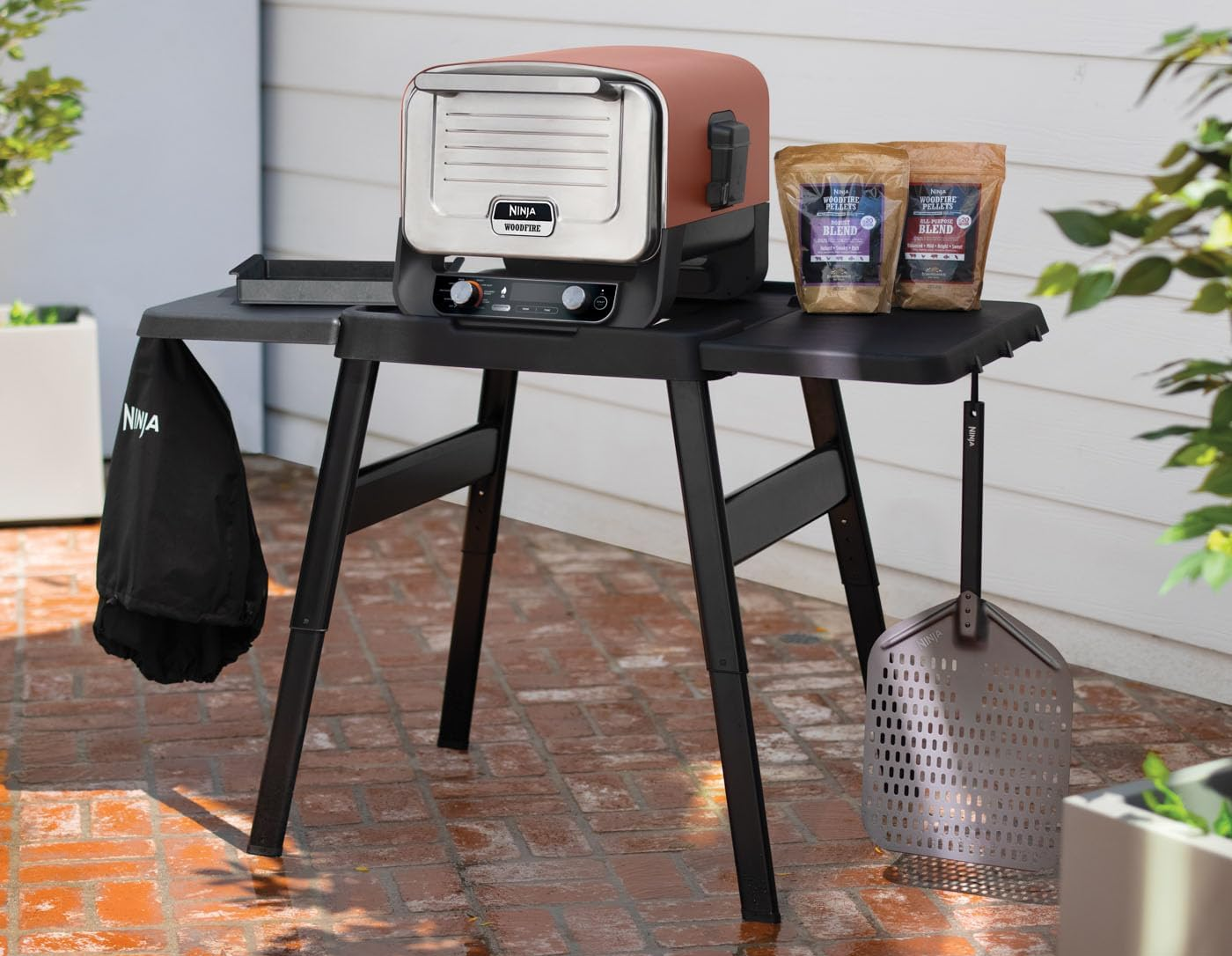 Egy Ninja Woodfire kültéri grill ételpellettel és kiegészítőkkel egy téglás teraszon lévő asztalon.