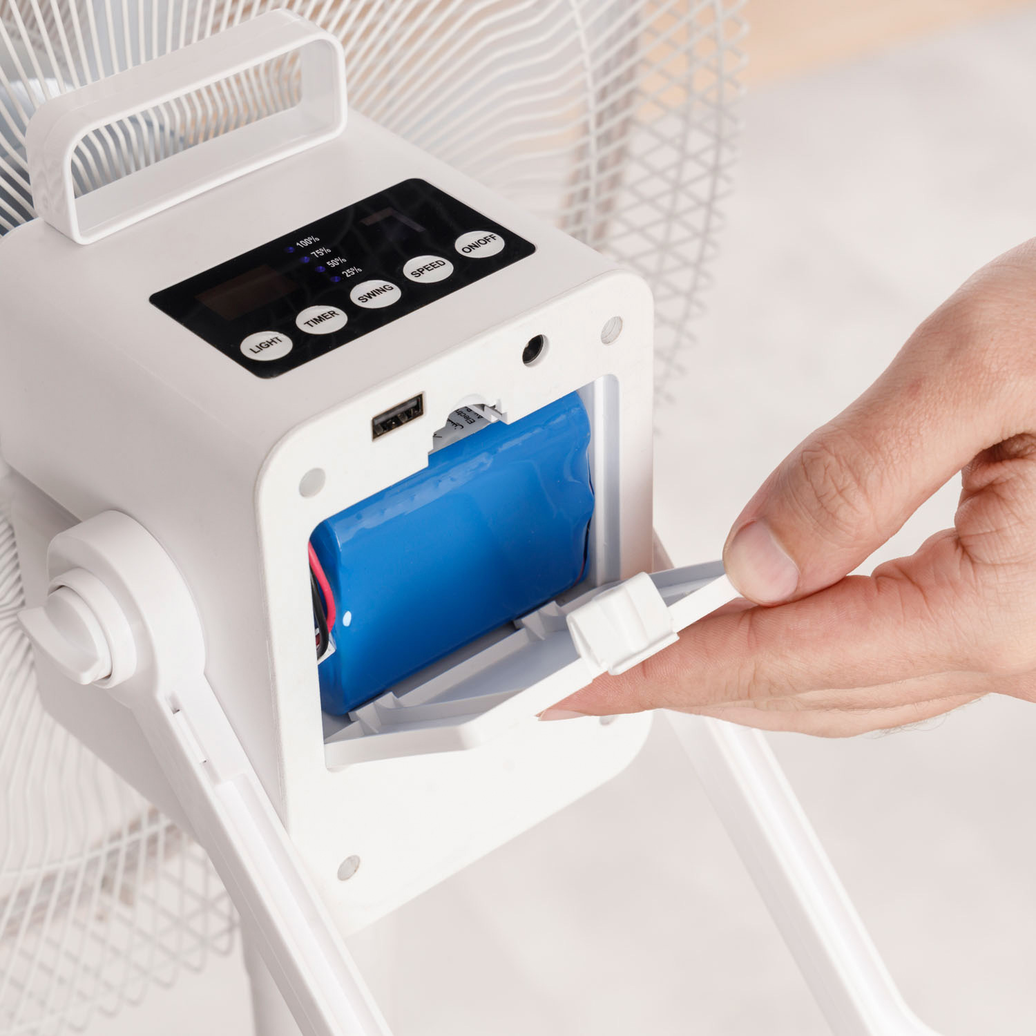 Un ventilador blanco con un panel de control y un compartimento de batería azul. Una mano abre el compartimento.