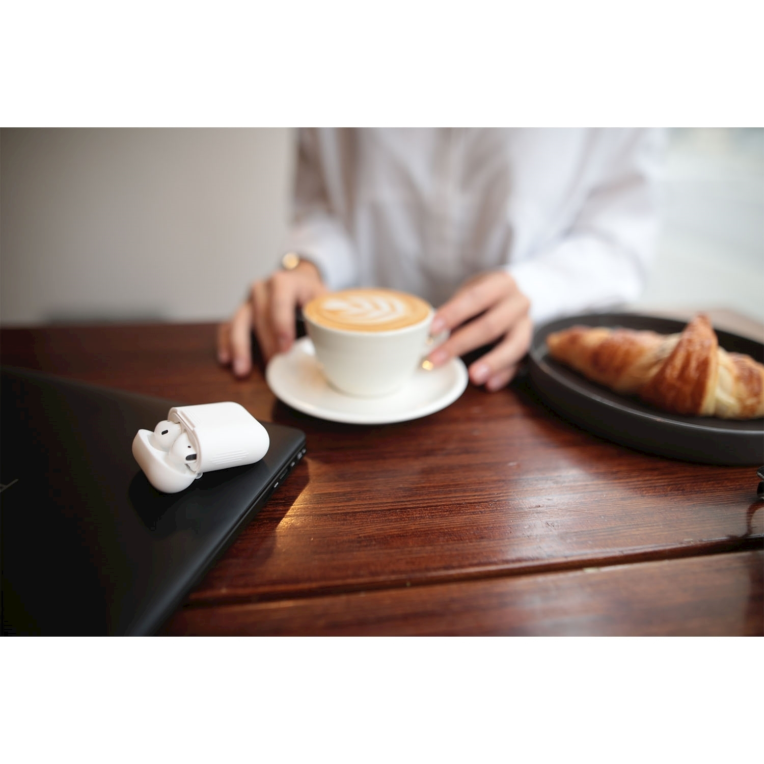 Una tazza di latte con un croissant su un piatto e auricolari bianchi aperti su un laptop nero.