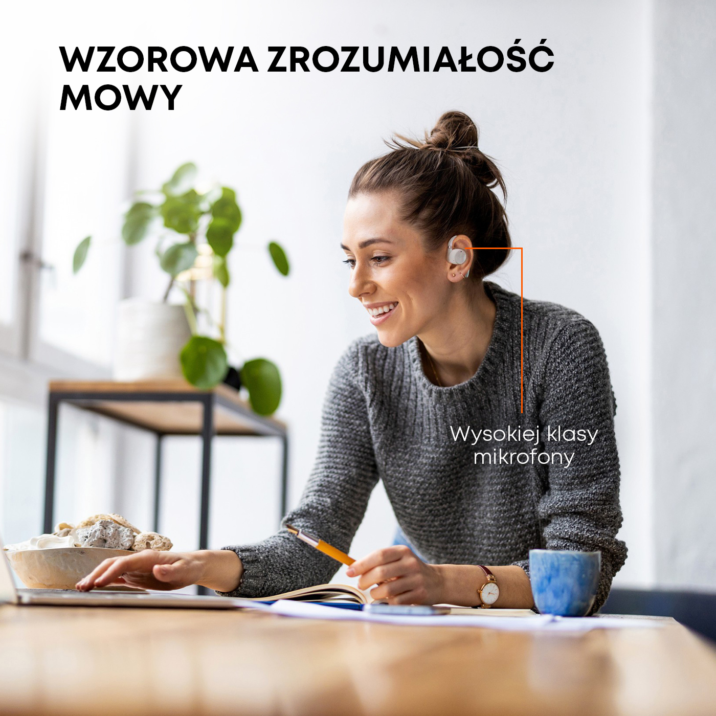 Kobieta w słuchawkach uśmiecha się podczas pracy na laptopie, z miską i kubkiem na biurku.