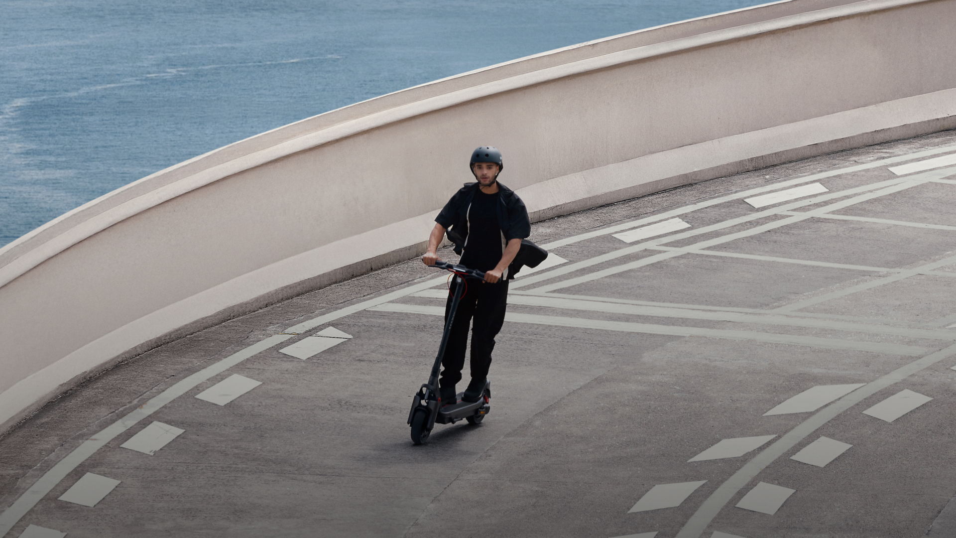 Mężczyzna w kasku jadący na skuterze elektrycznym po krętej drodze z widokiem na morze.