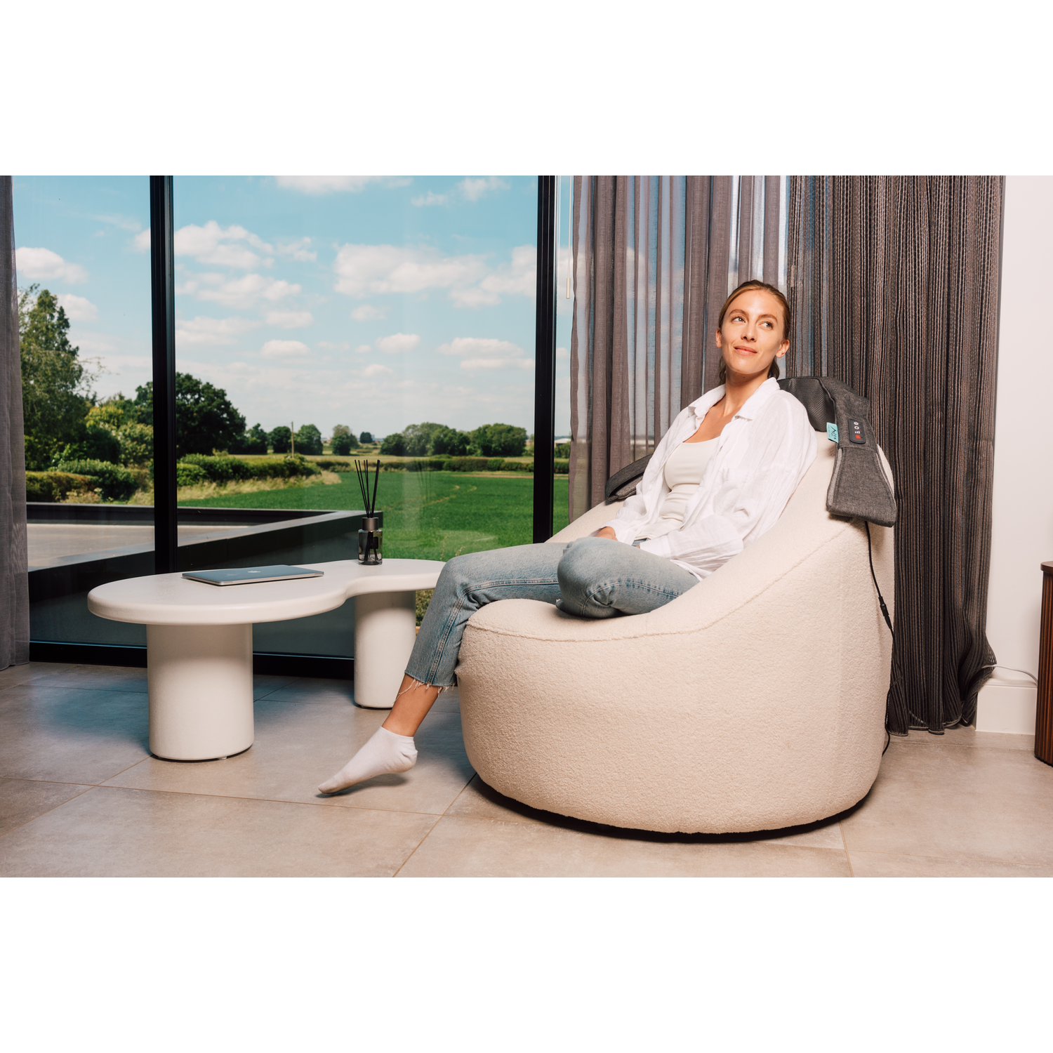 A woman relaxes in a white beanbag chair, wearing jeans and a shirt. A tablet and diffuser are near.