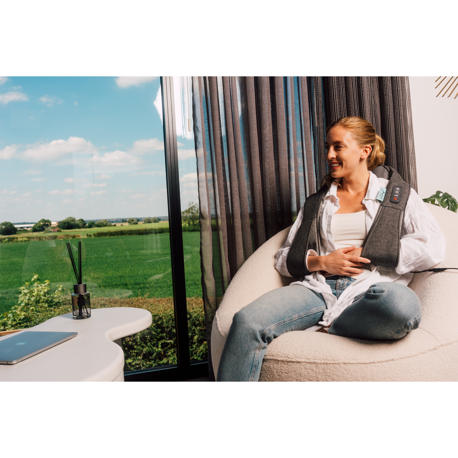 A woman wearing a grey massage device sits on a chair near a window, smiling. The background has a view.