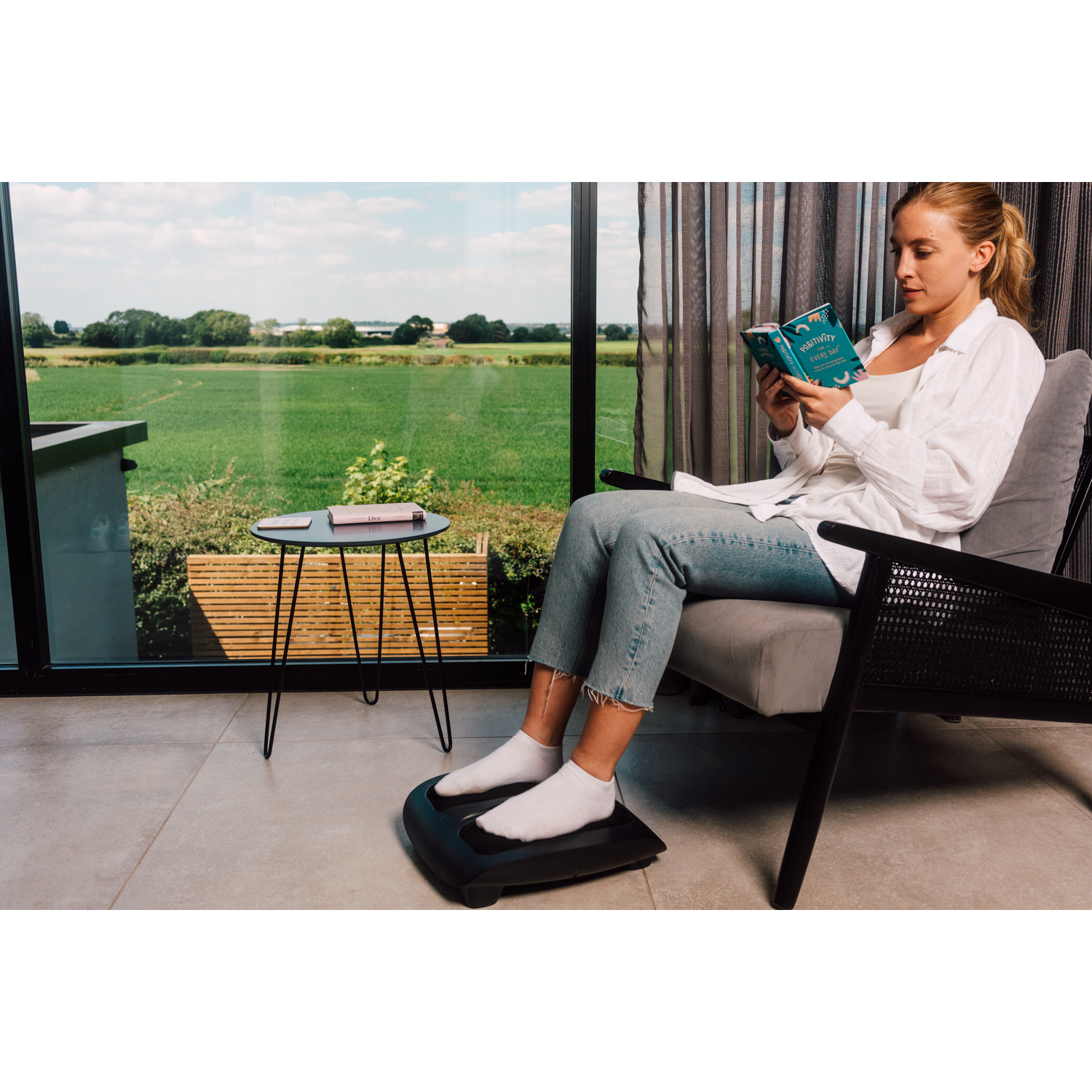 A woman sits reading a book, her feet resting on a black foot massager near a window.