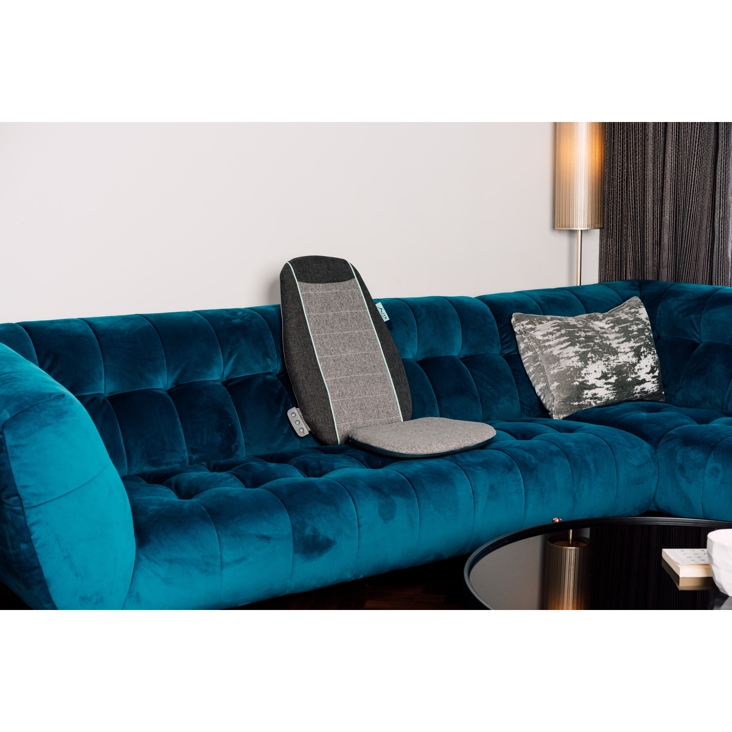 Un canapé en velours sarcelle avec un coussin de soutien dorsal. Une table en verre foncé et une lampe.