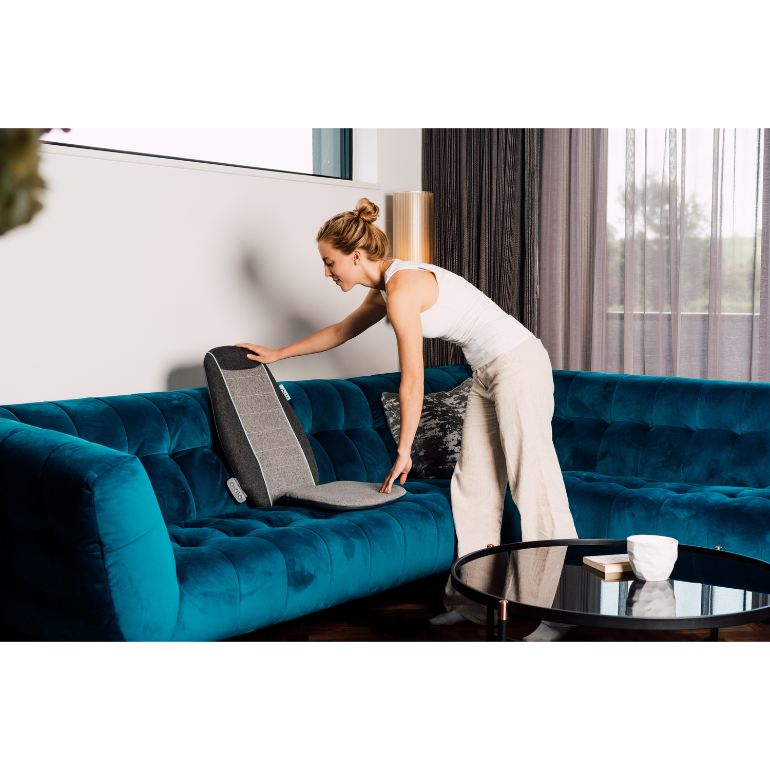 Une femme plaçant un coussin de soutien lombaire gris et noir sur un canapé turquoise.