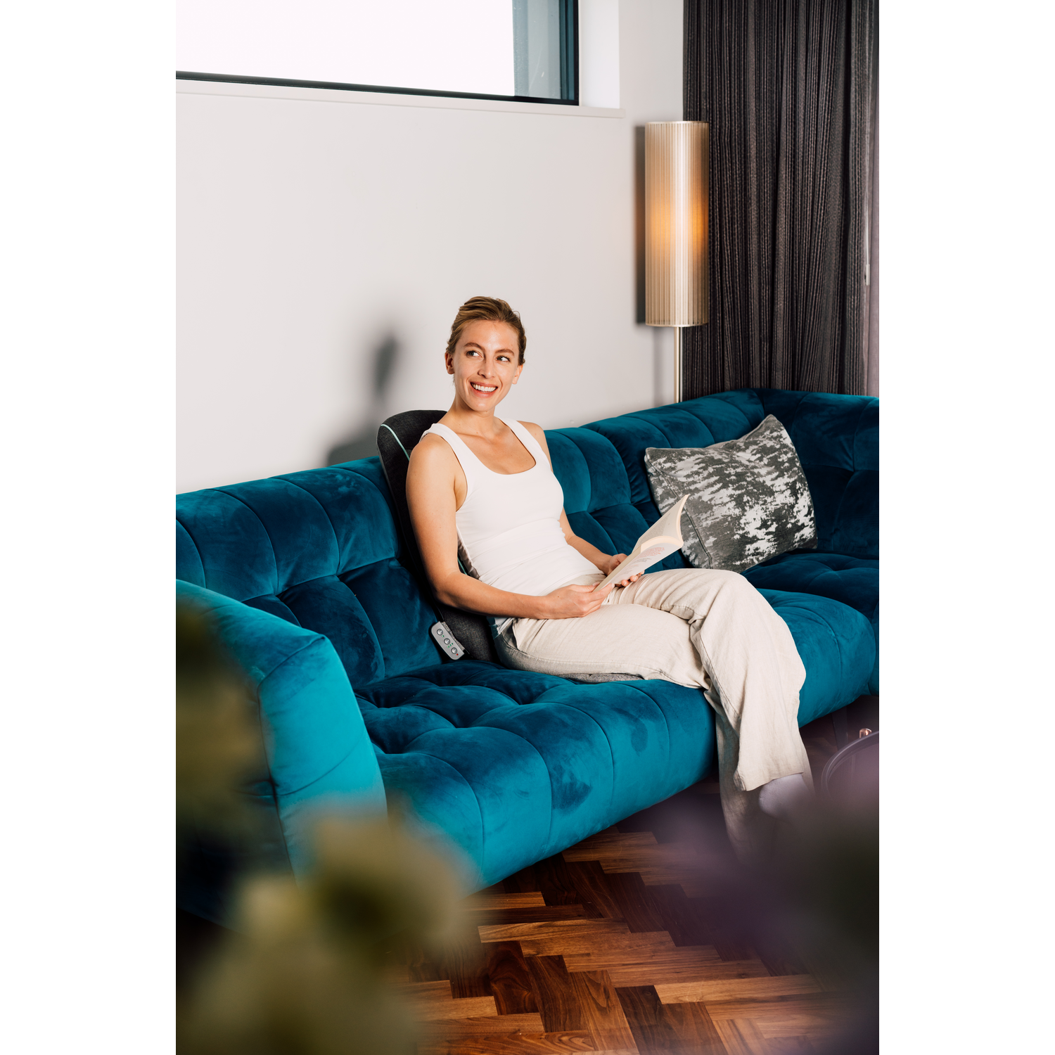 Femme sur un canapé turquoise avec un oreiller et un masseur de cou, lisant, lumière vive.