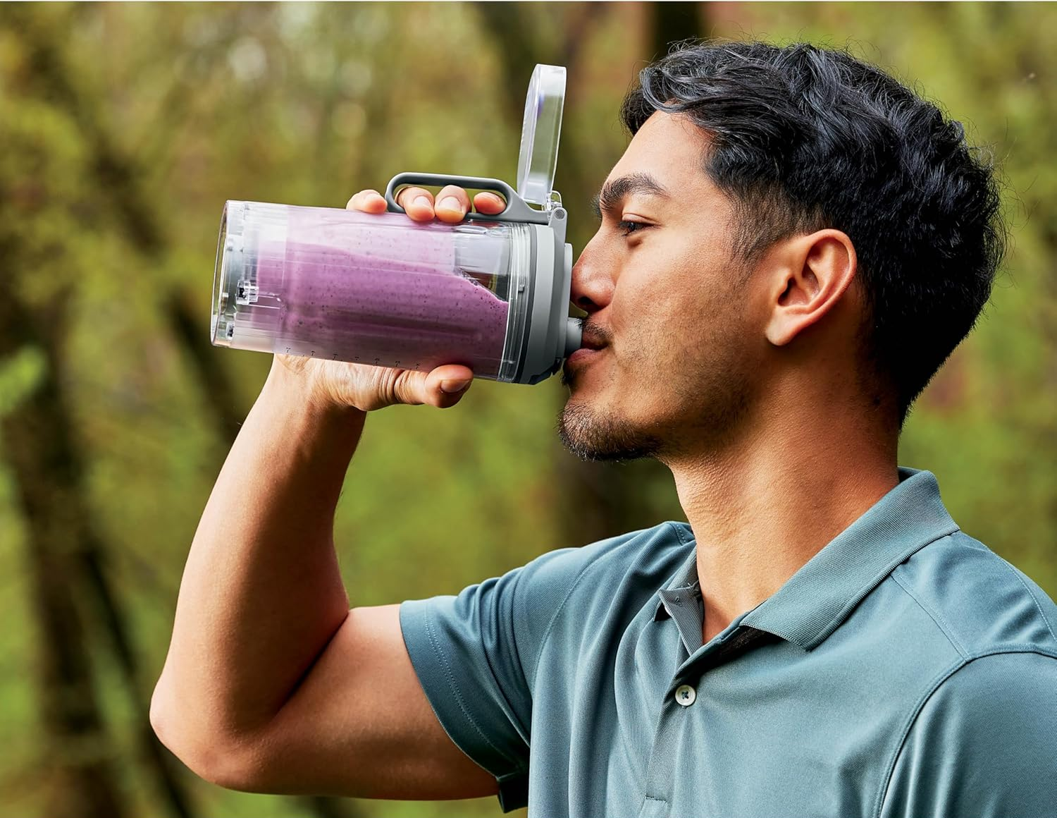 Férfi lila smoothie-s palackból iszik. Zöld háttér fákkal. Palack átlátszó.