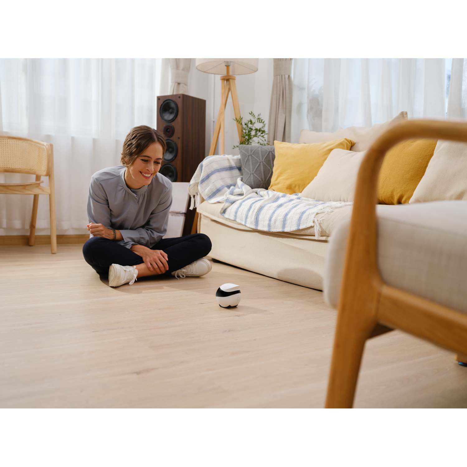 A woman is sitting on the floor with a small, black and white robotic ball. The background is a room.