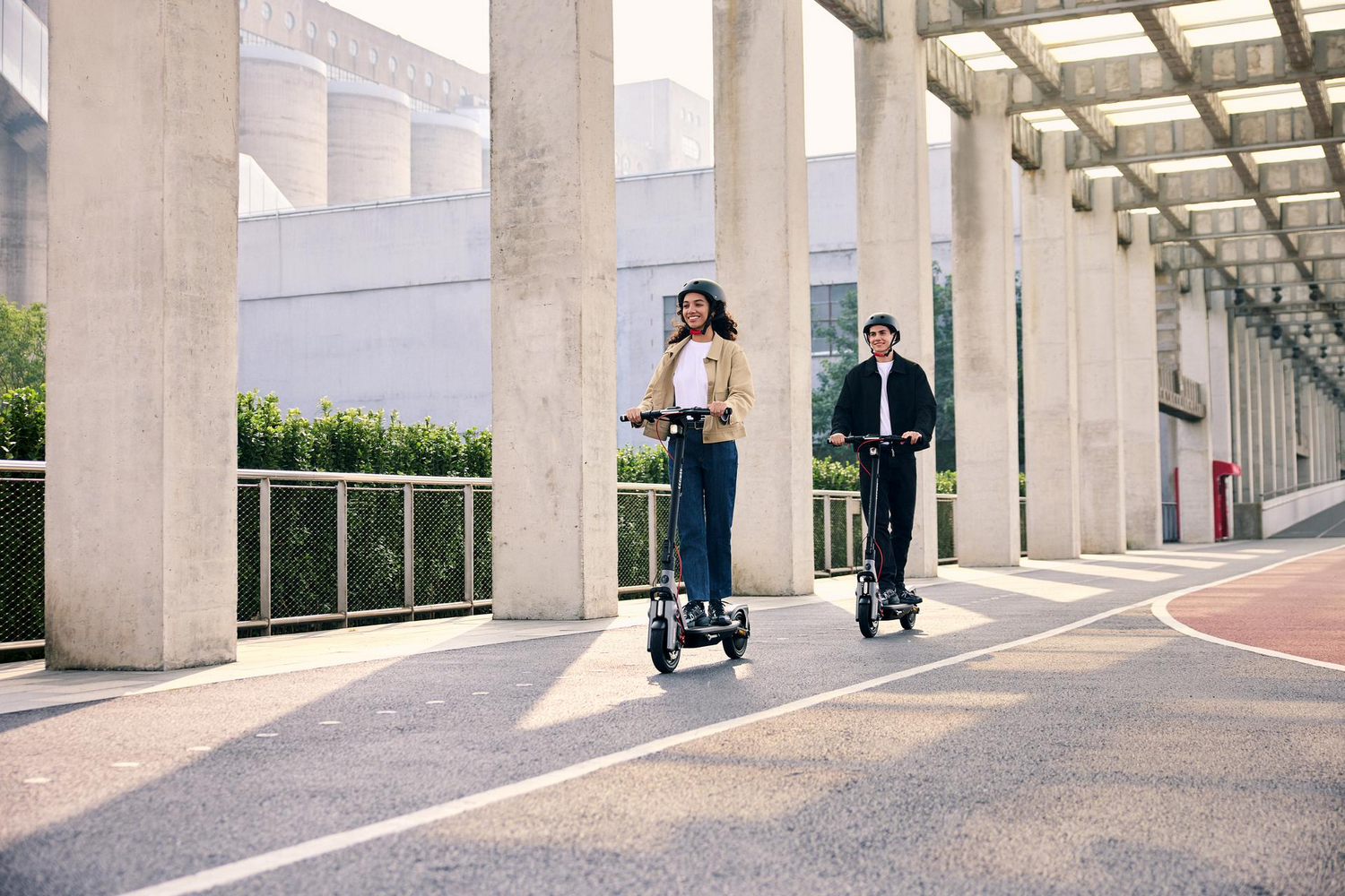 Dos personas en patinetes montan en un camino bajo una pasarela cubierta. Edificios en el fondo.