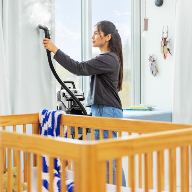 Kobieta parująca ścianę urządzeniem czyszczącym w pobliżu łóżeczka.