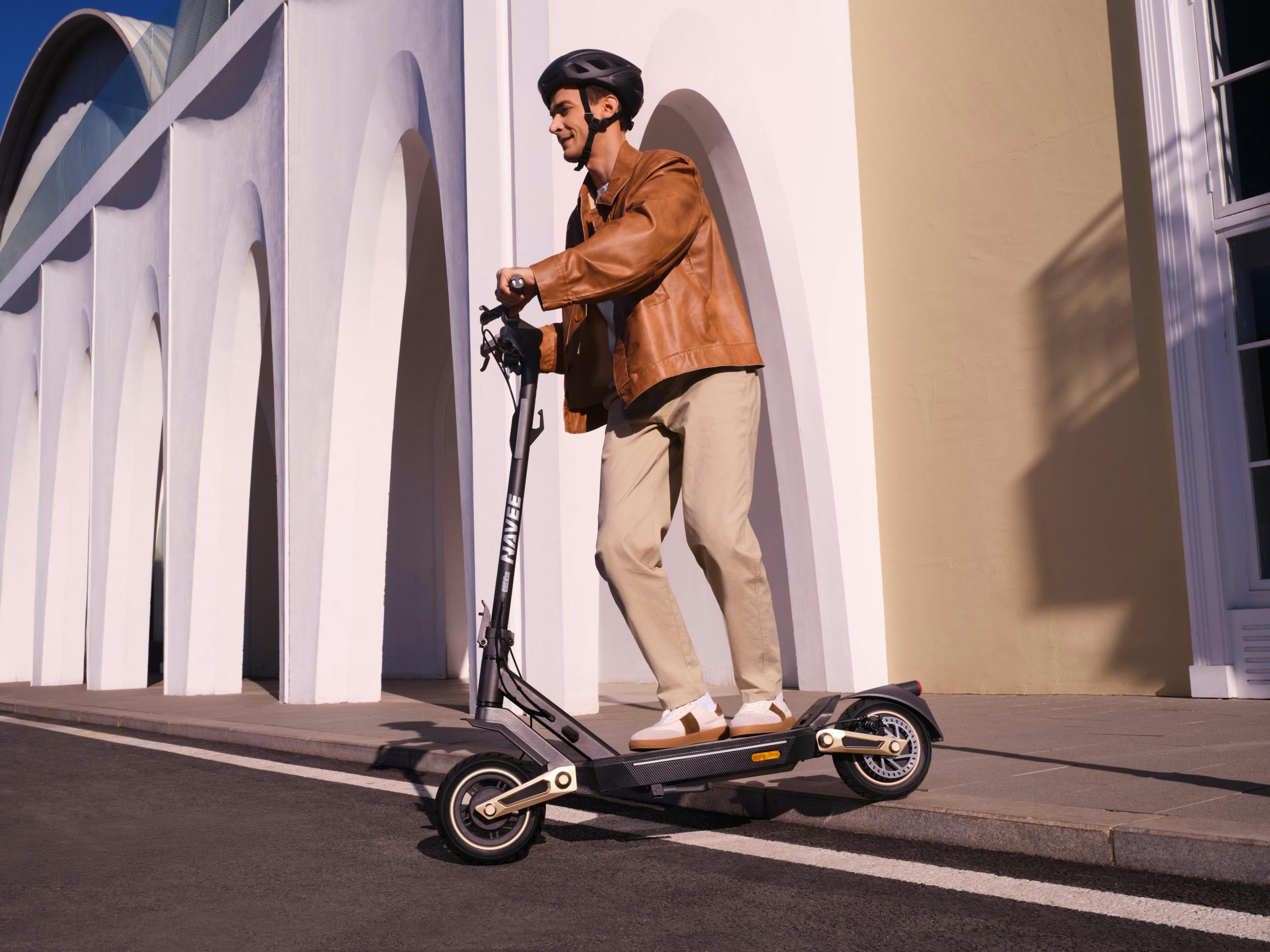 Ein Mann fährt mit einem Elektroroller auf der Straße. Er trägt Helm und braune Jacke.