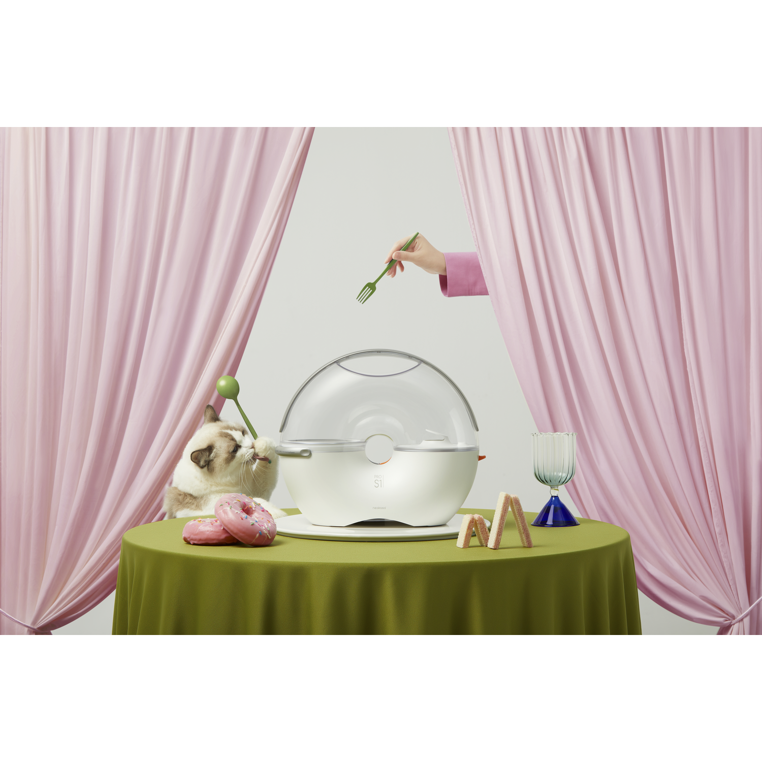A cat eats a donut near food and a table set-up with a pink curtain in the background.