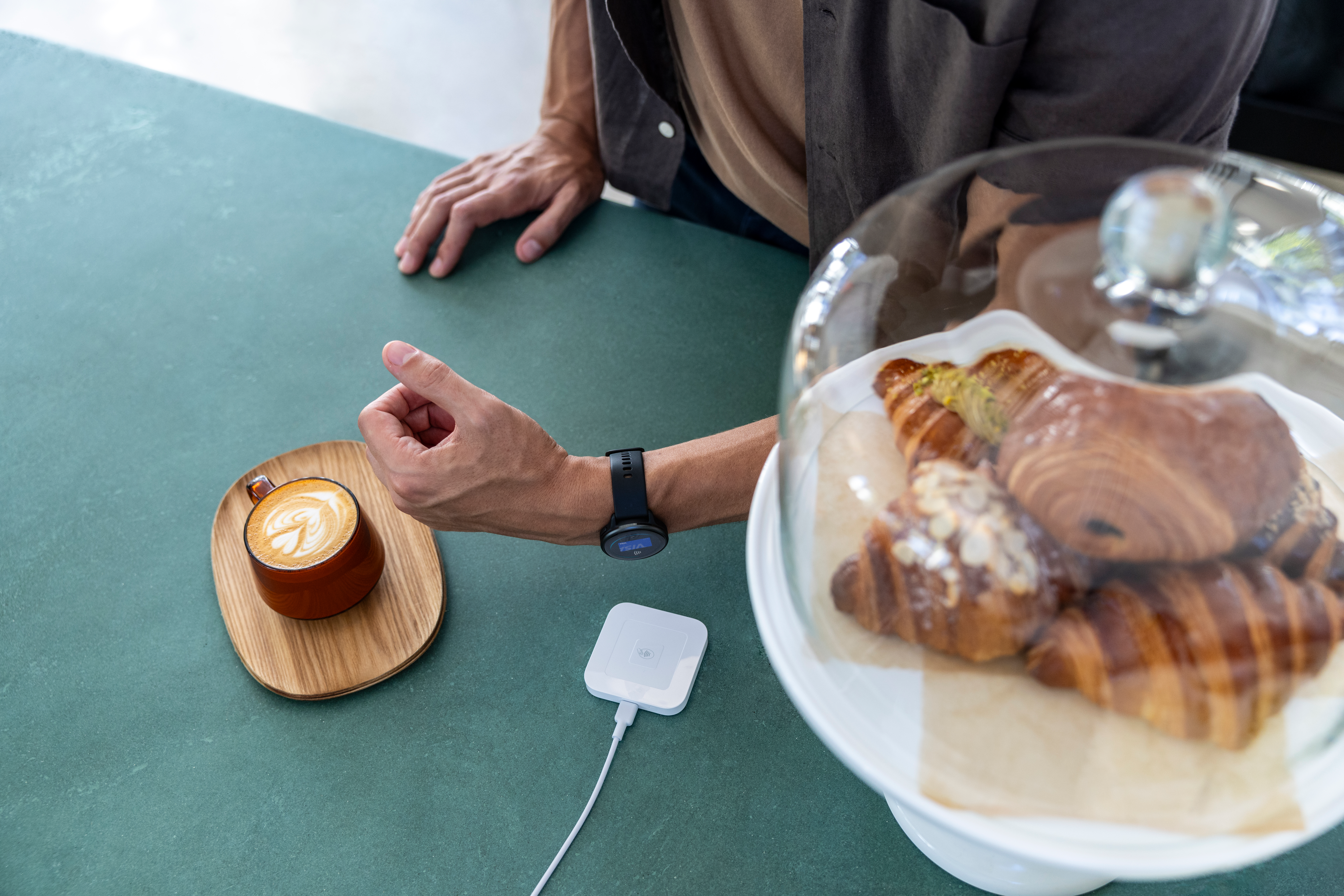 Personne avec une smartwatch et du café sur un plateau en bois. Croissants sous cloche en verre.
