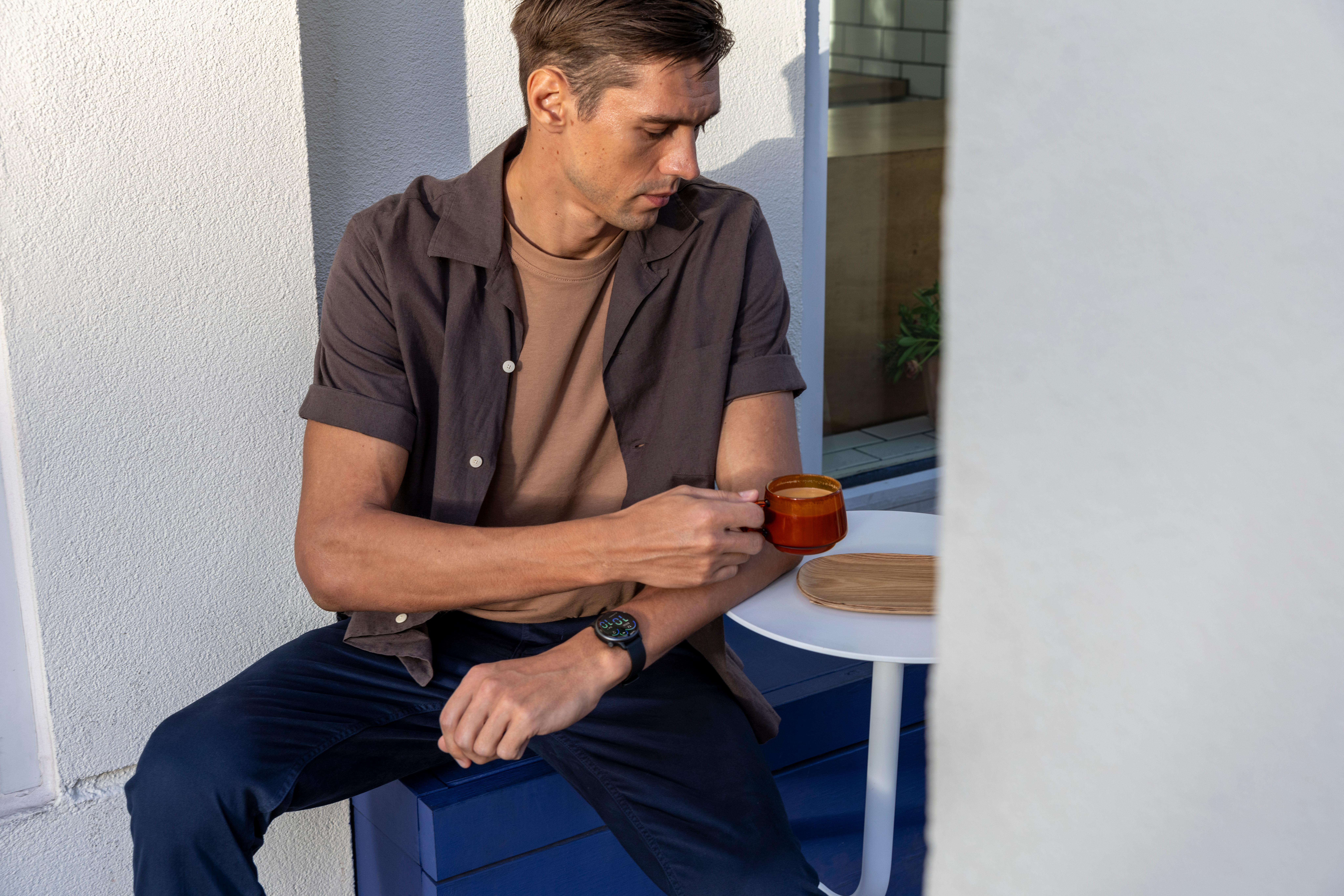 Un homme assis à une table, tenant une tasse de café. Il porte une chemise marron et un pantalon bleu.