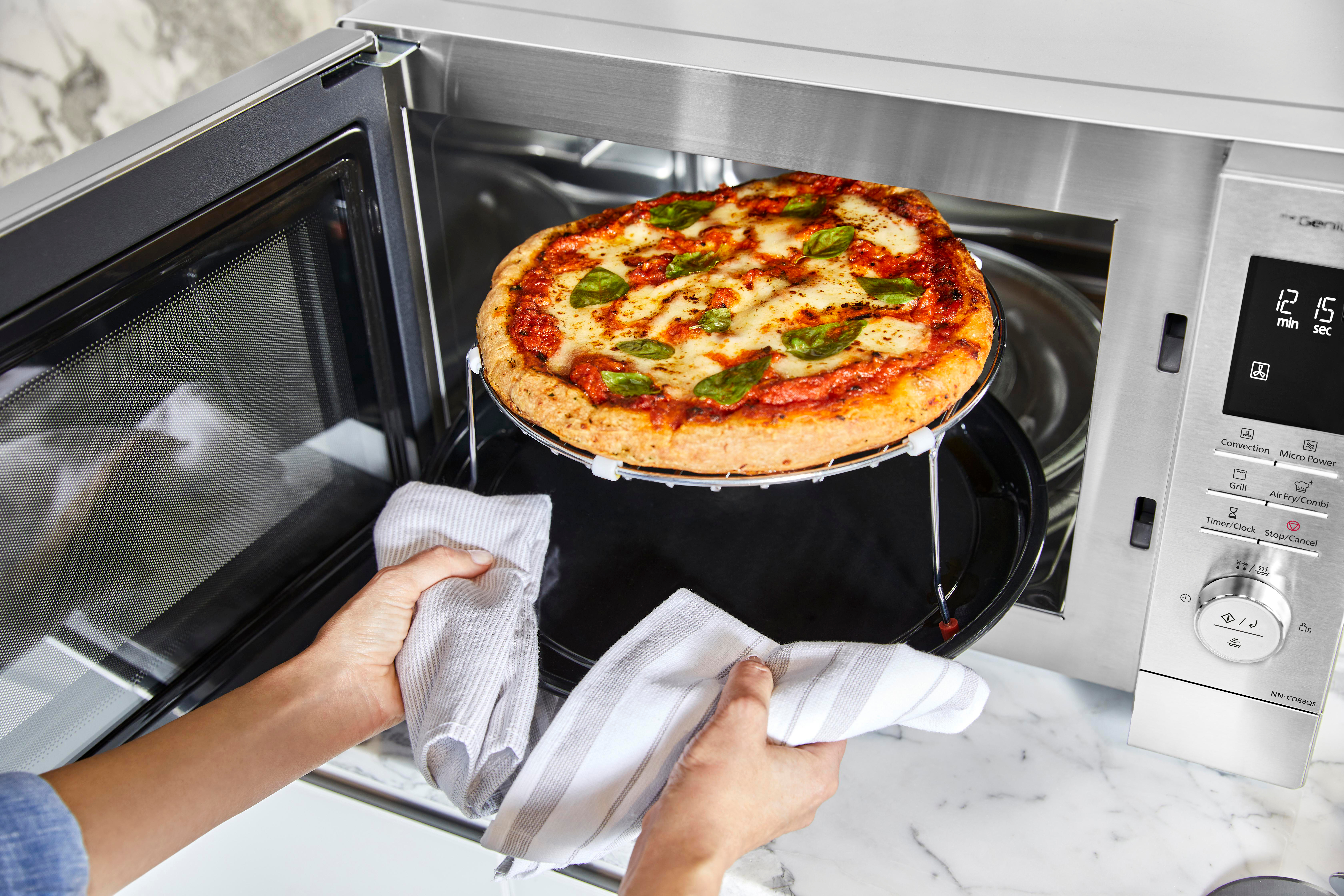 Eine Pizza auf einem Rost in einer Mikrowelle, Hände halten Tücher.