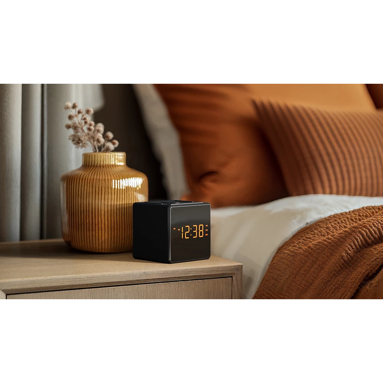 A black cube clock displays 12:38 on a wooden nightstand with a vase of dried flowers, next to a bed with orange bedding.