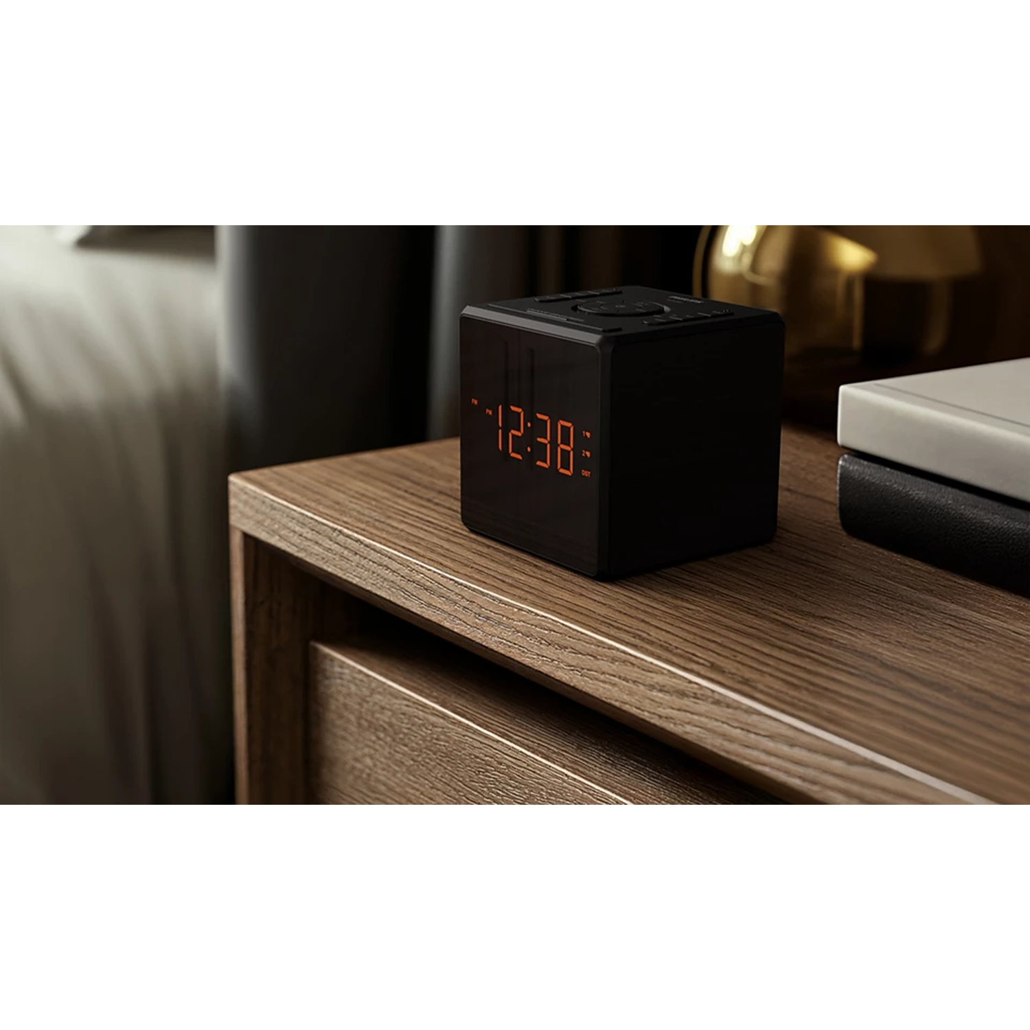 A black cube-shaped digital clock displays 12:38 on a wooden nightstand. Books are beside the clock, and a blurred bed is in the background.