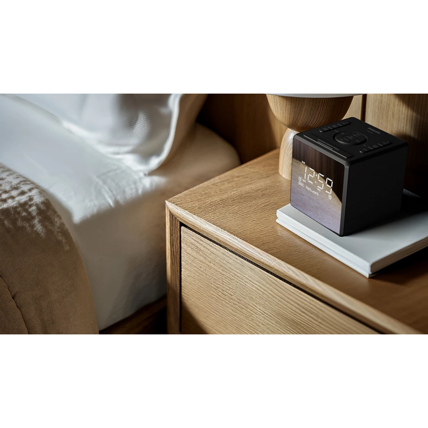 A black digital clock displays 12:59 on a bedside table. Beside it is a bed with white sheets and a wooden nightstand.