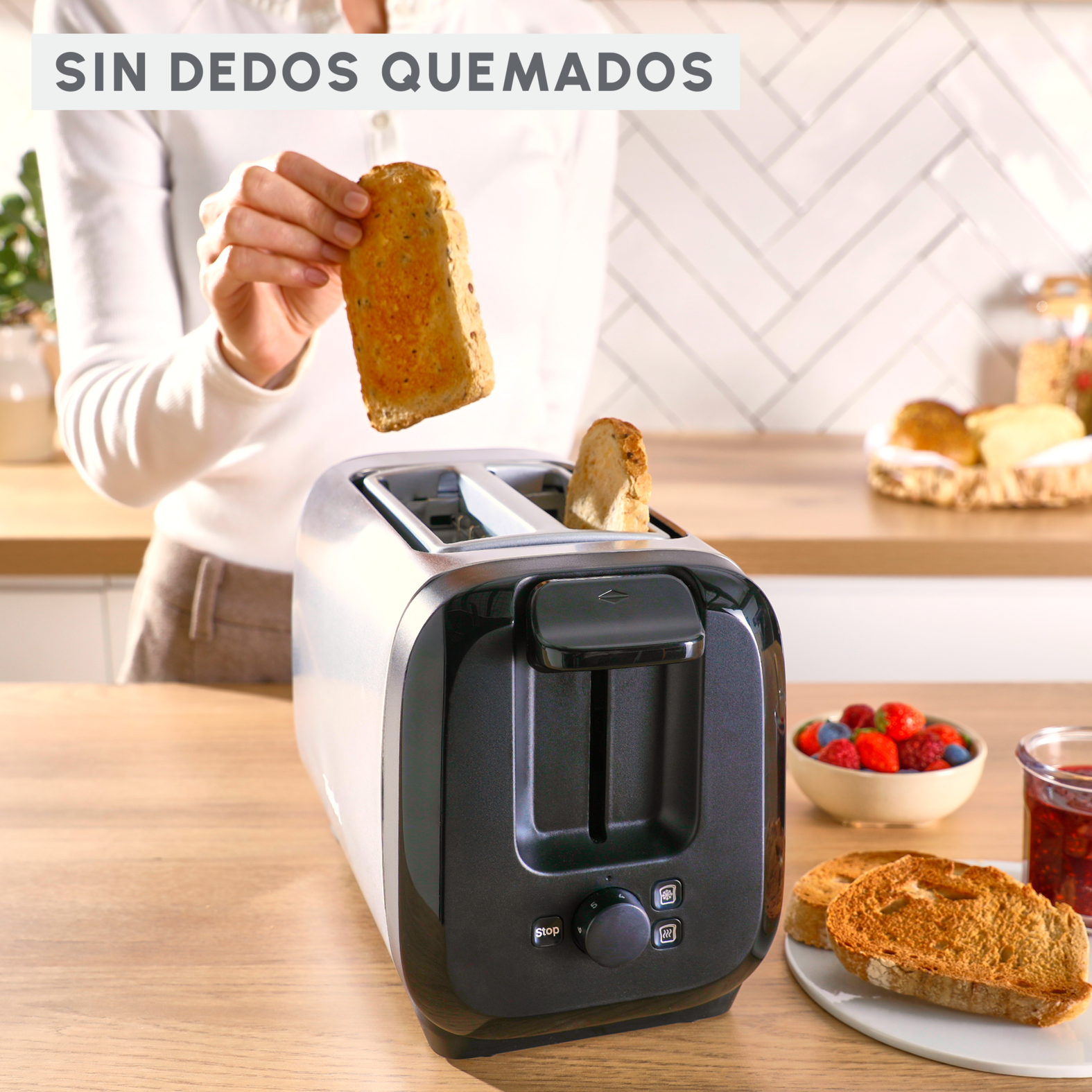 Una mujer sosteniendo una tostada sobre una tostadora, con un cuenco de bayas y mermelada en la mesa.